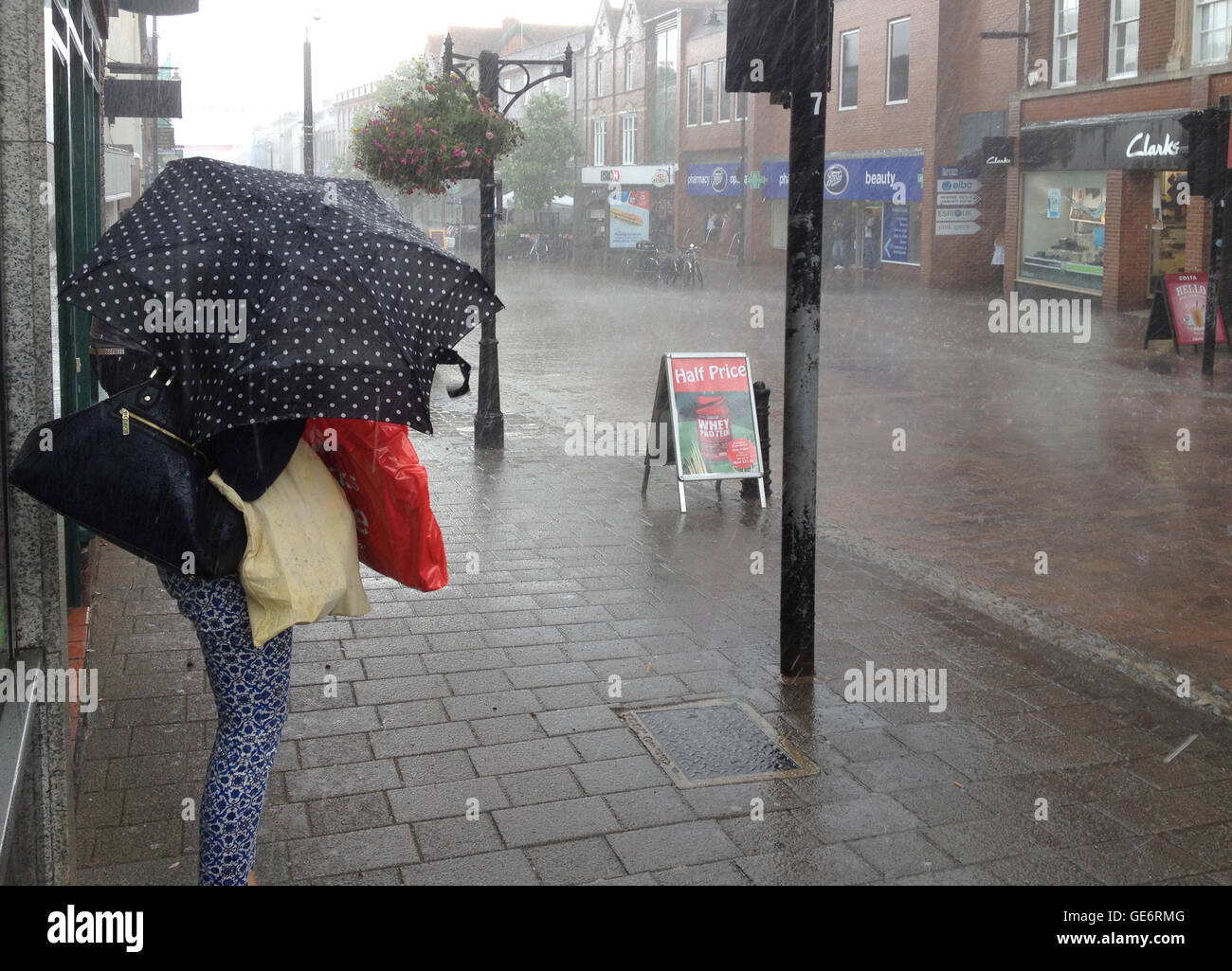 Una donna cercando rifugio per se stessa e per le sue borse della spesa dalla pioggia torrenziale con un piccolo ombrello nella cittadina di Newbury, Berkshire, Regno Unito Foto Stock