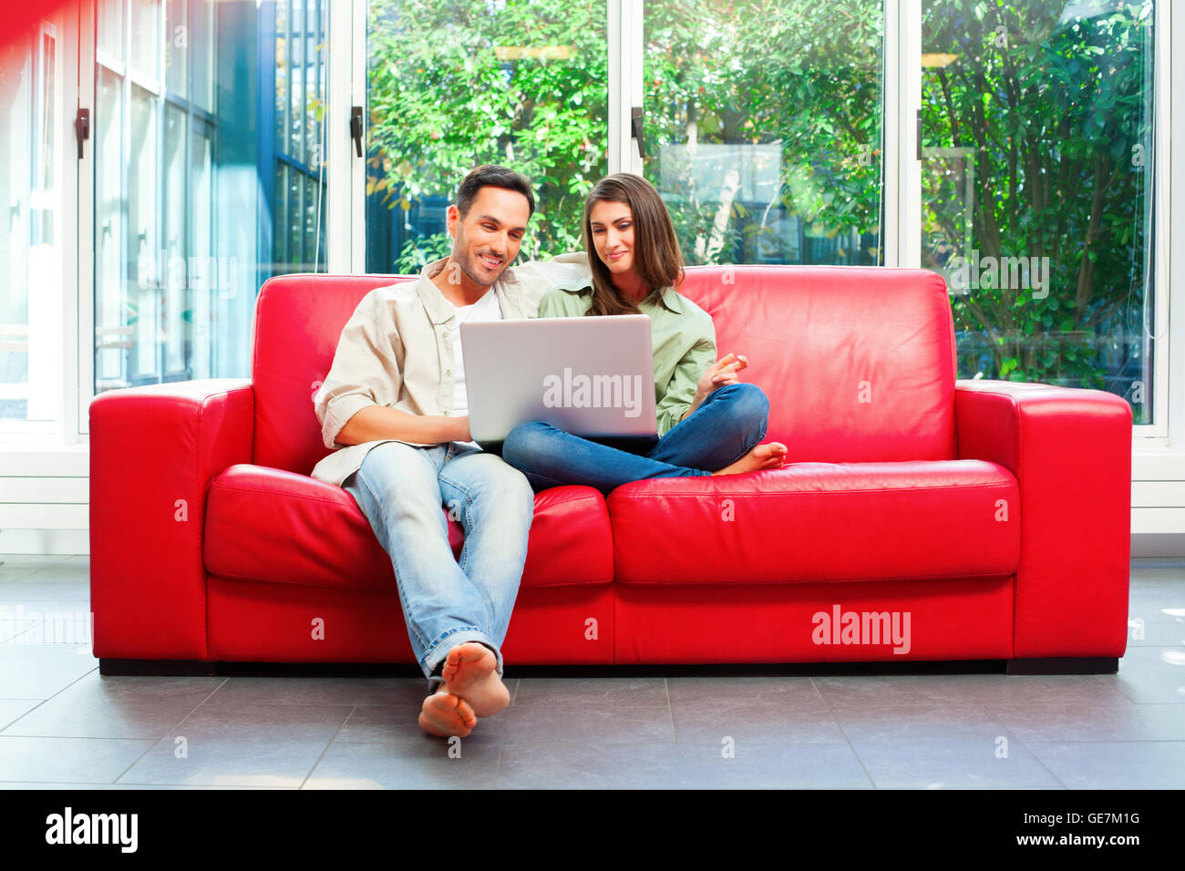 Per tutta la lunghezza della coppia giovane utilizzando laptop. Felice maschio e femmina dei partner sono seduti sul divano rosso. Essi sono in casuals a casa. Foto Stock