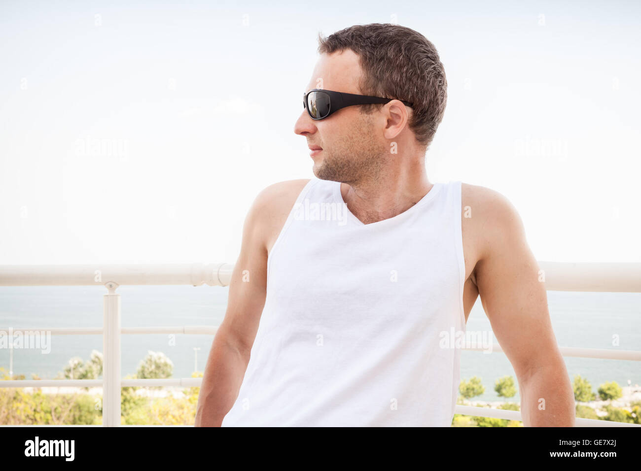 Giovani caucasici sportivo Uomo in camicia bianca e occhiali da sole. Dehor Estivo ritratto con mare su uno sfondo Foto Stock