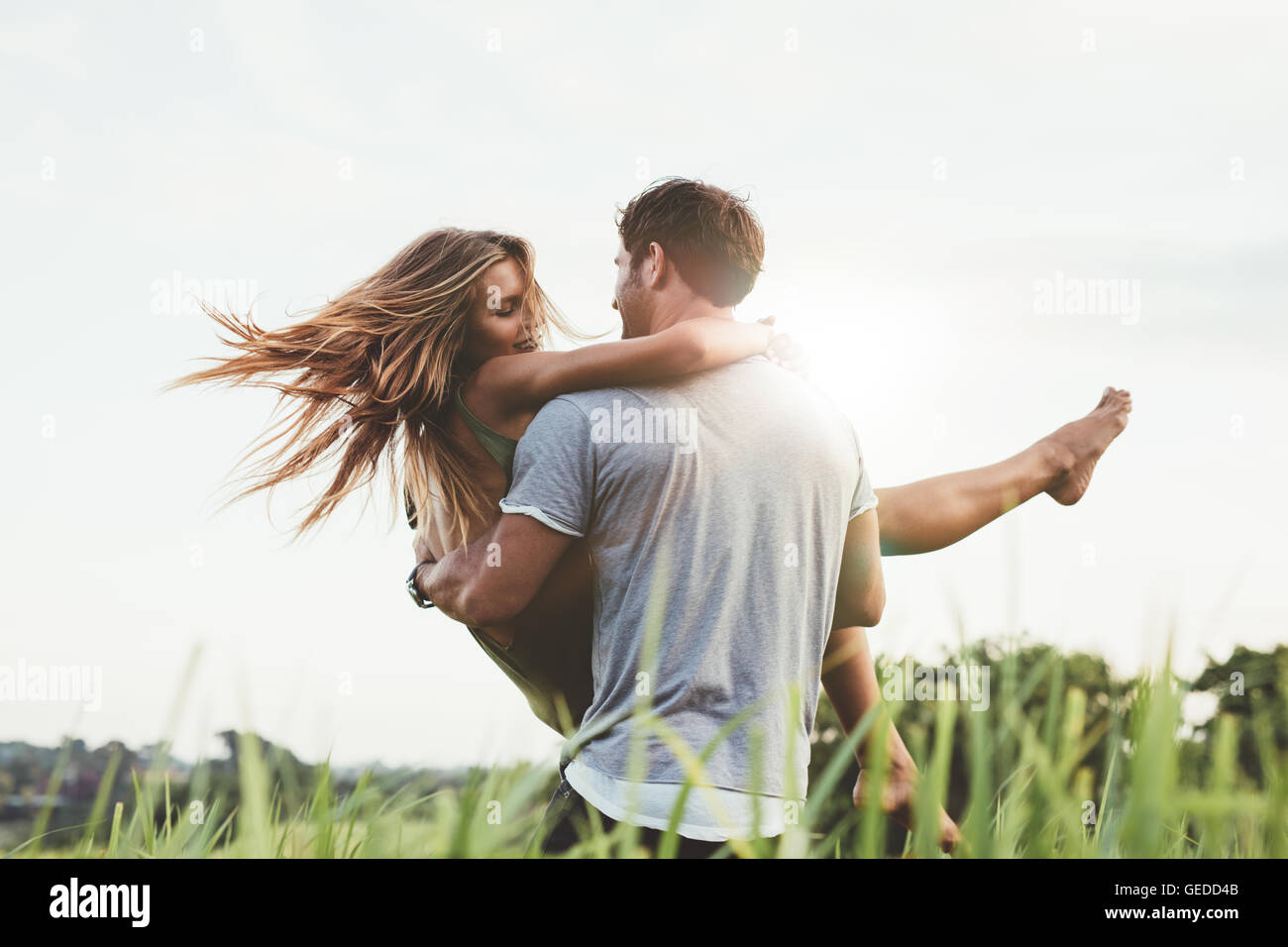 Colpo di giovane portando la sua fidanzata a braccia sul campo in erba. Matura per divertirsi in natura in un giorno di estate. Foto Stock