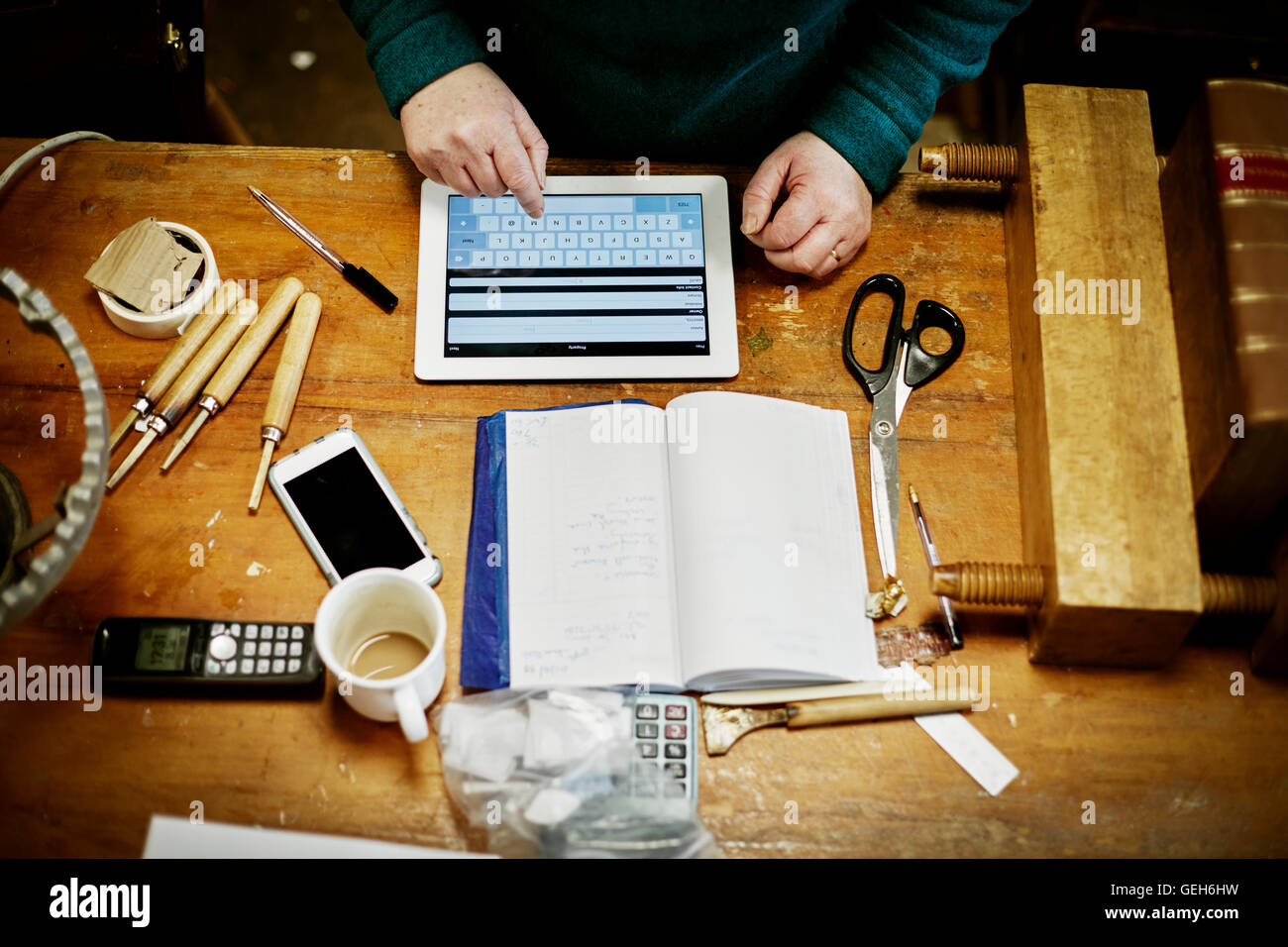 Una persona seduta su un banco da lavoro, utilizzando una tavoletta digitale. Gli strumenti forbici e uno smart phone sul banco. Foto Stock