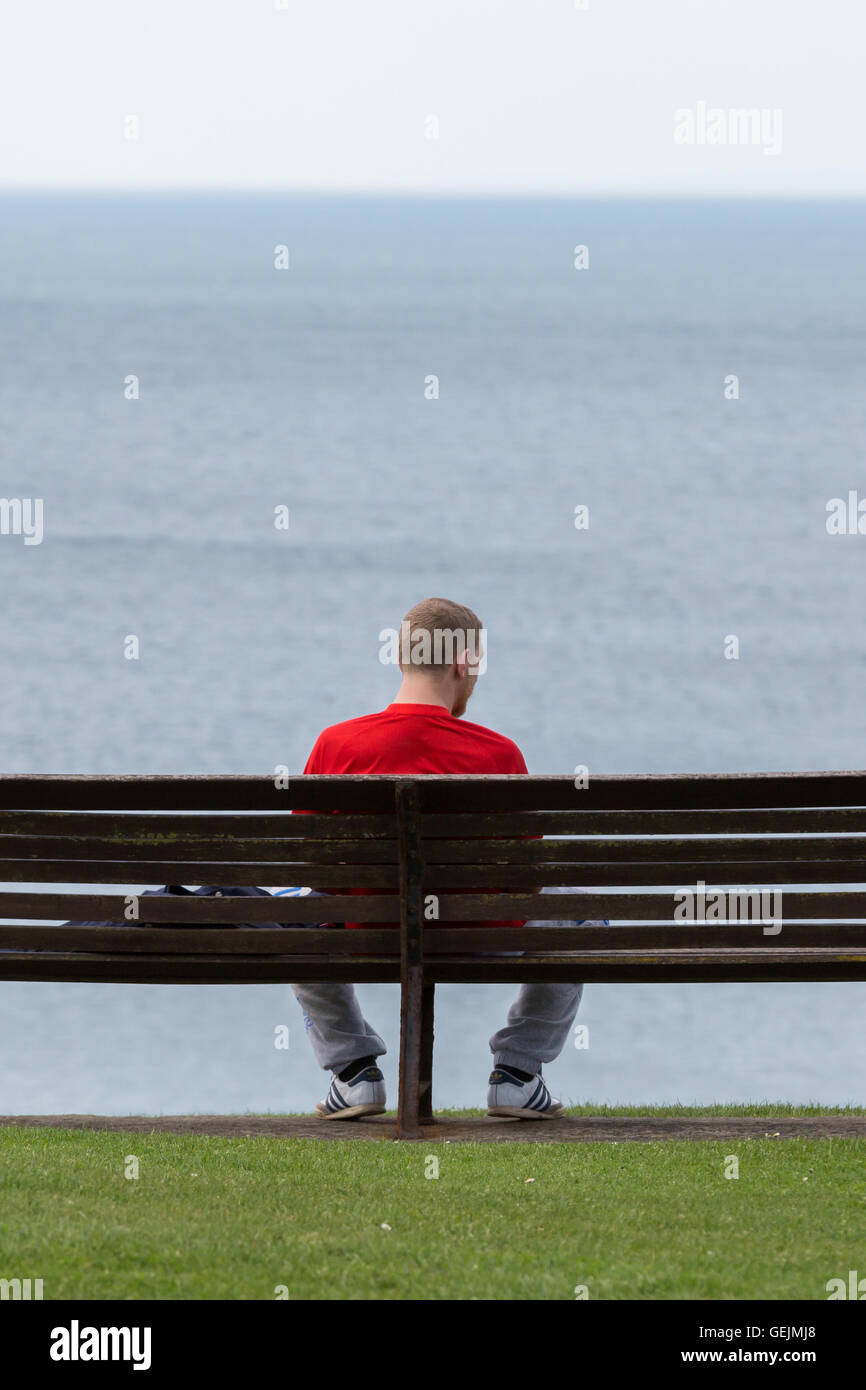 Un uomo seduto su una panchina nel parco che guarda al mare Foto Stock
