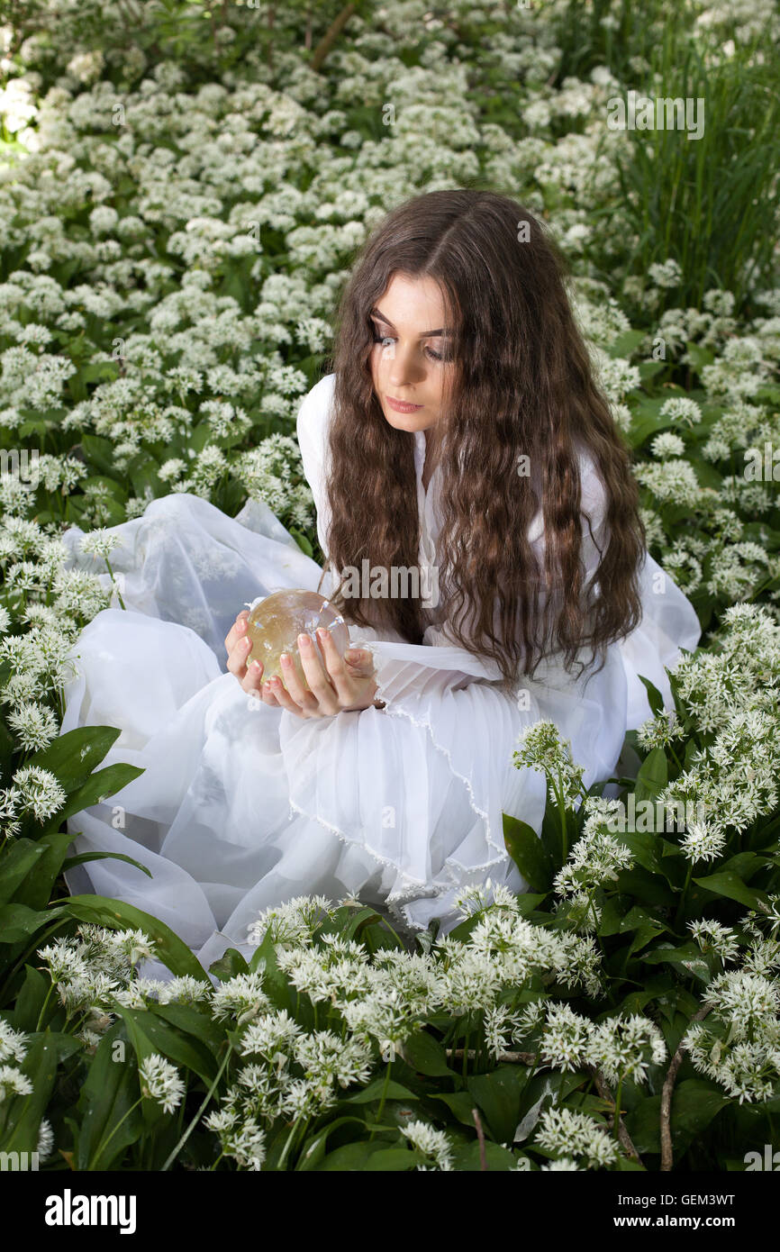 Giovane e bella donna che indossa un abito bianco in una foresta guardando nella sfera di cristallo Foto Stock