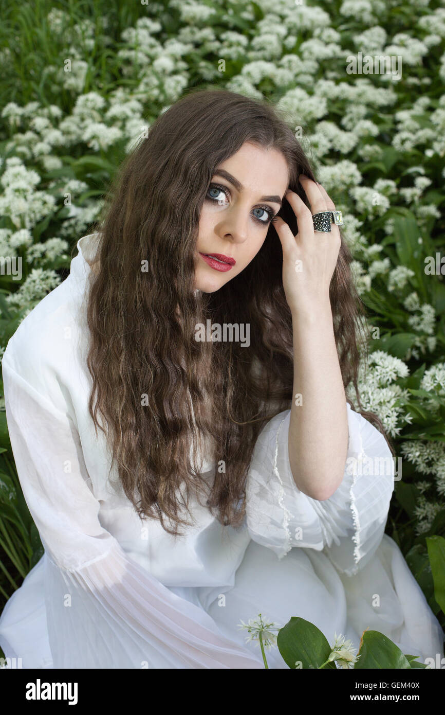 Bella donna che indossa un lungo abito bianco seduto tra i fiori bianchi tenendo la mano al suo viso Foto Stock