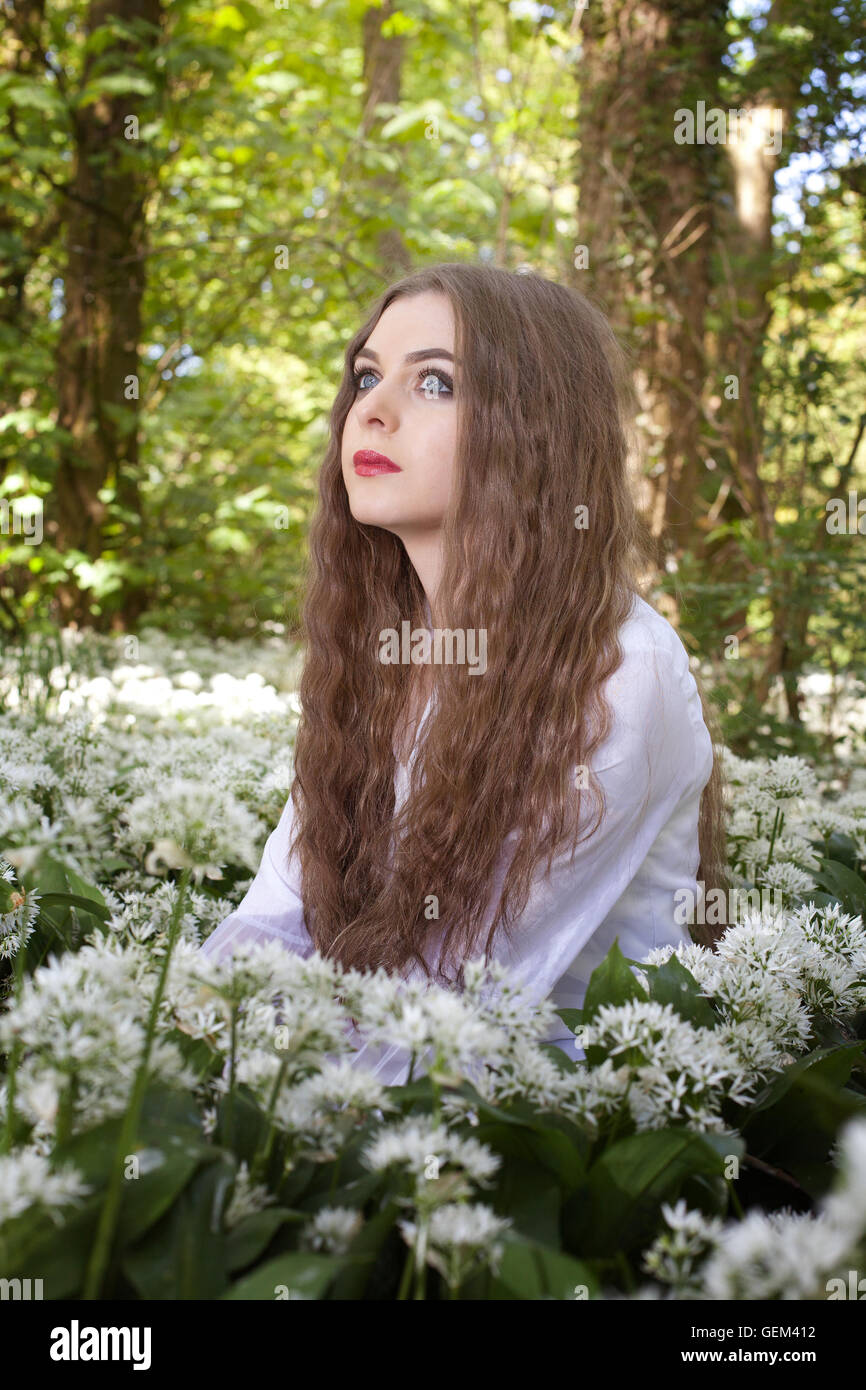 Bella donna che indossa un lungo abito bianco in seduta fiori bianchi guardando in lontananza Foto Stock