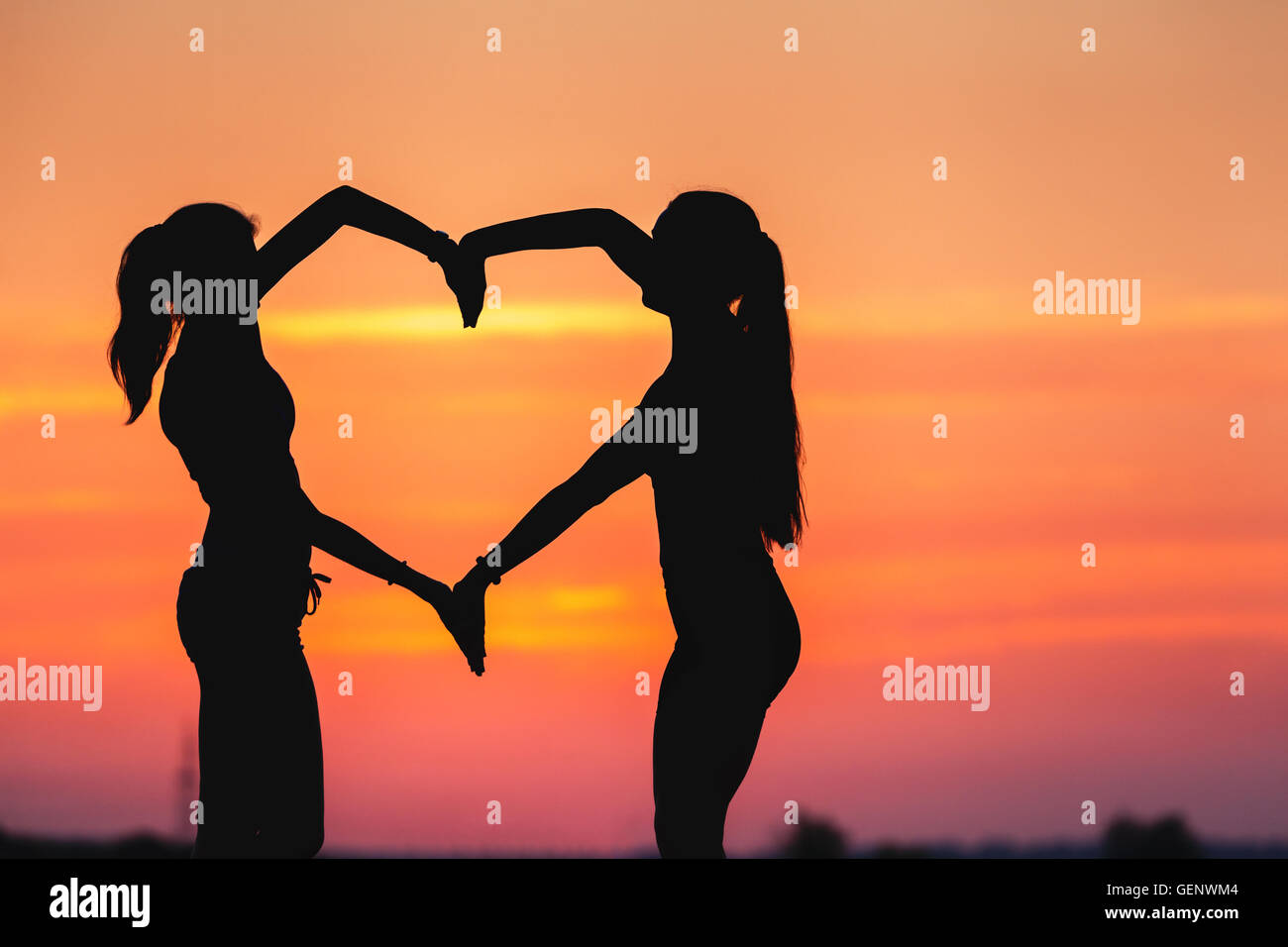 Paesaggio con silhouette di giovani donne sportivo tenendo le mani in forma di cuore su uno sfondo di cielo colorato al tramonto in summ Foto Stock