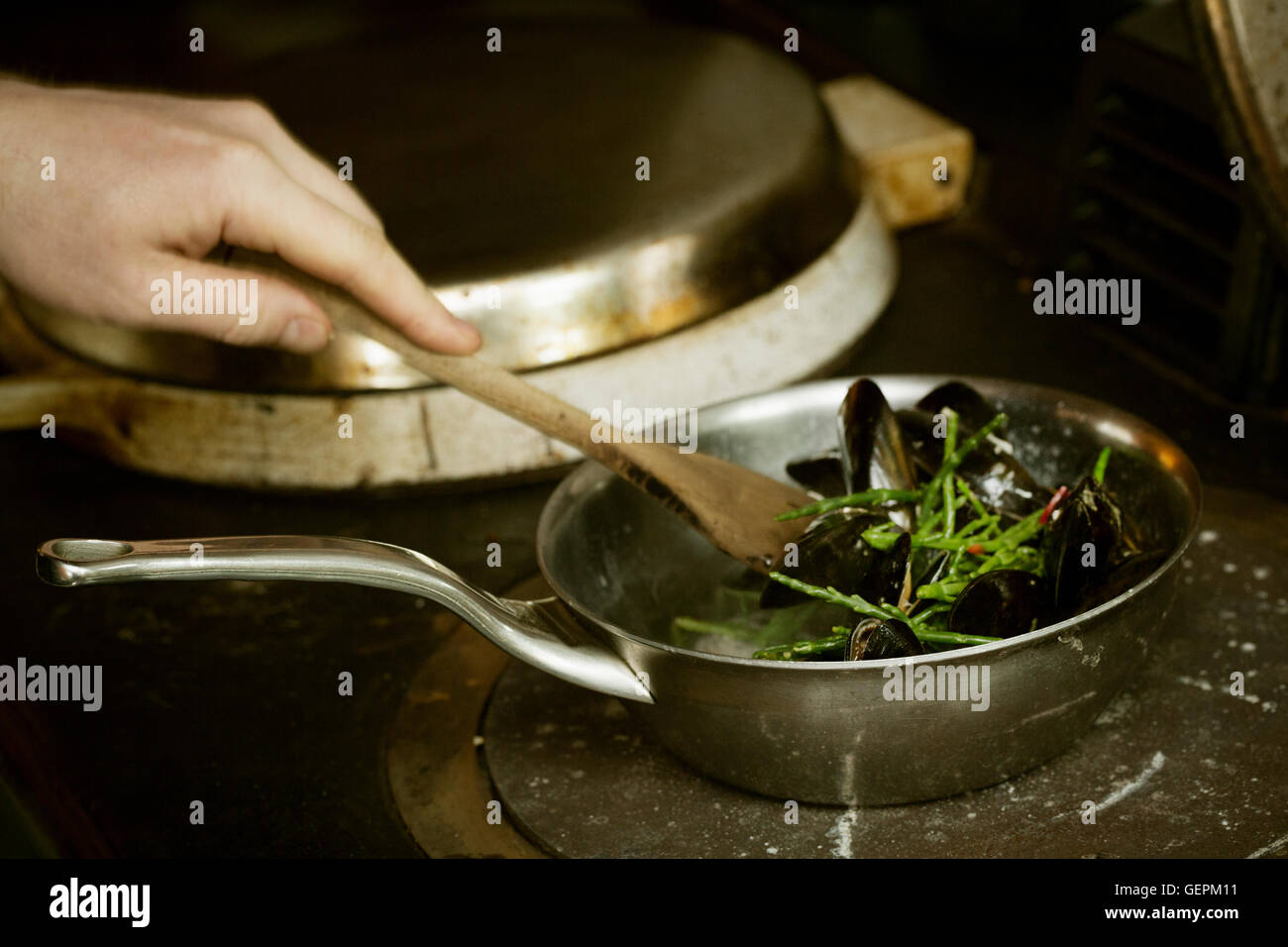 Chef nero di agitazione di cozze e samphire in un tegame su un fornello. Foto Stock