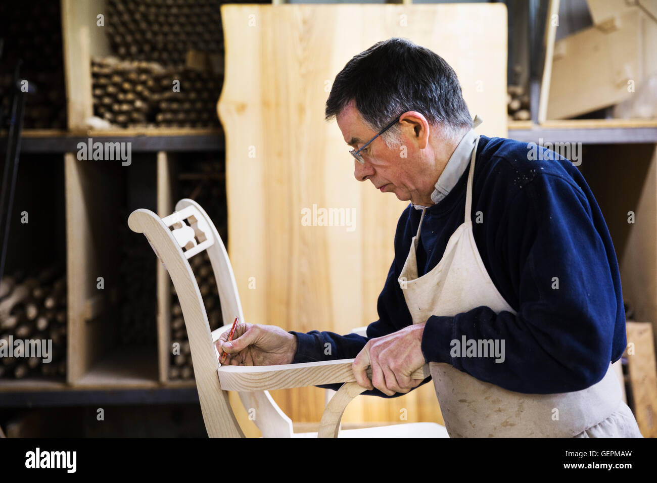 Uomo in piedi in un laboratorio di falegnameria, lavorando su una sedia in legno la marcatura del giunto del bracciolo con una matita. Foto Stock