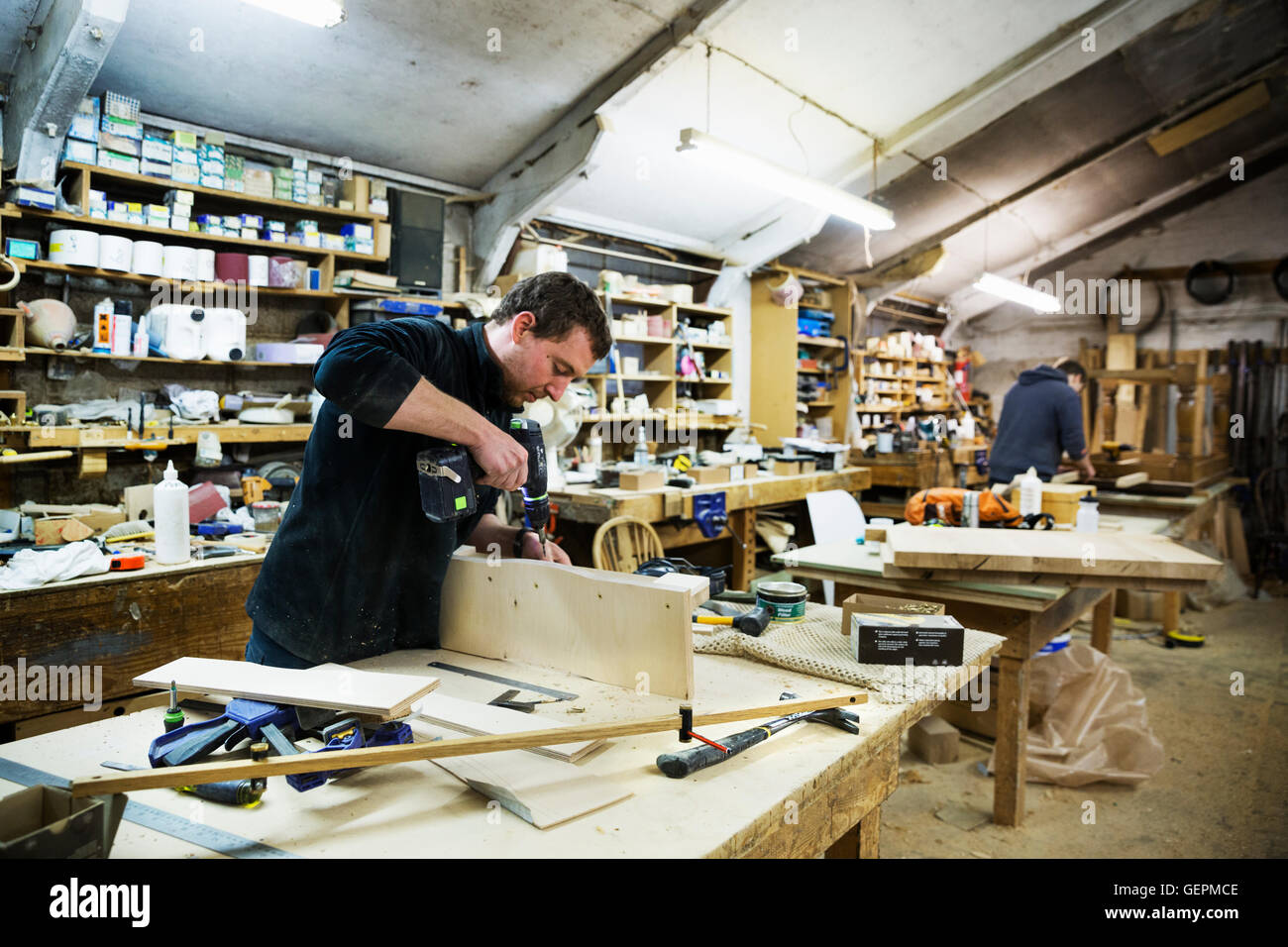 Uomo in piedi in corrispondenza di un banco di lavoro in un laboratorio di falegnameria, lavorando su un pezzo di legno, utilizzando un trapano. Foto Stock
