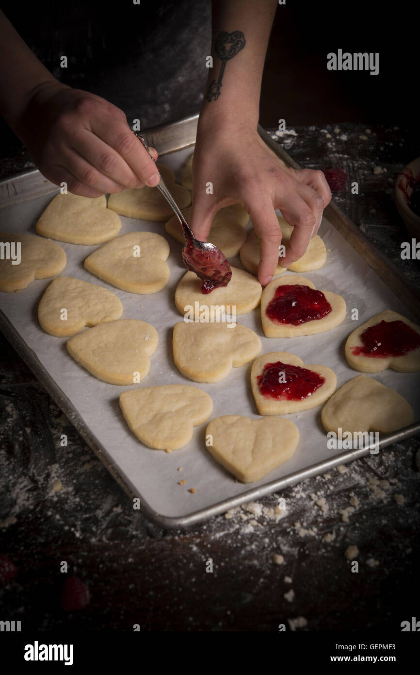 Il giorno di San Valentino la cottura, donna diffusione di confettura di lamponi sul cuore biscotti di forma. Foto Stock