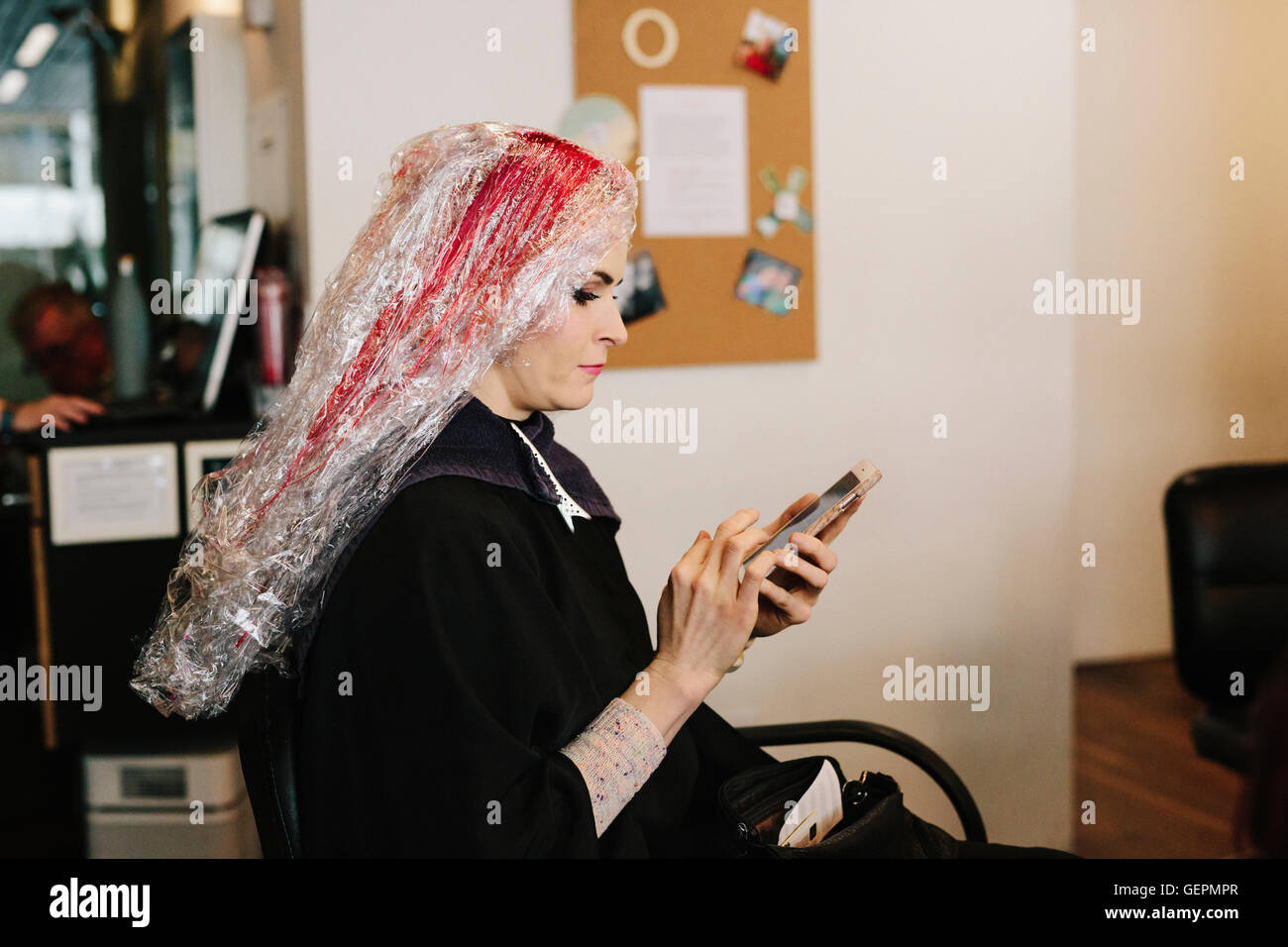 Una donna in un parrucchiere avente un colore di capelli trattamento, utilizzando un telefono intelligente. Foto Stock
