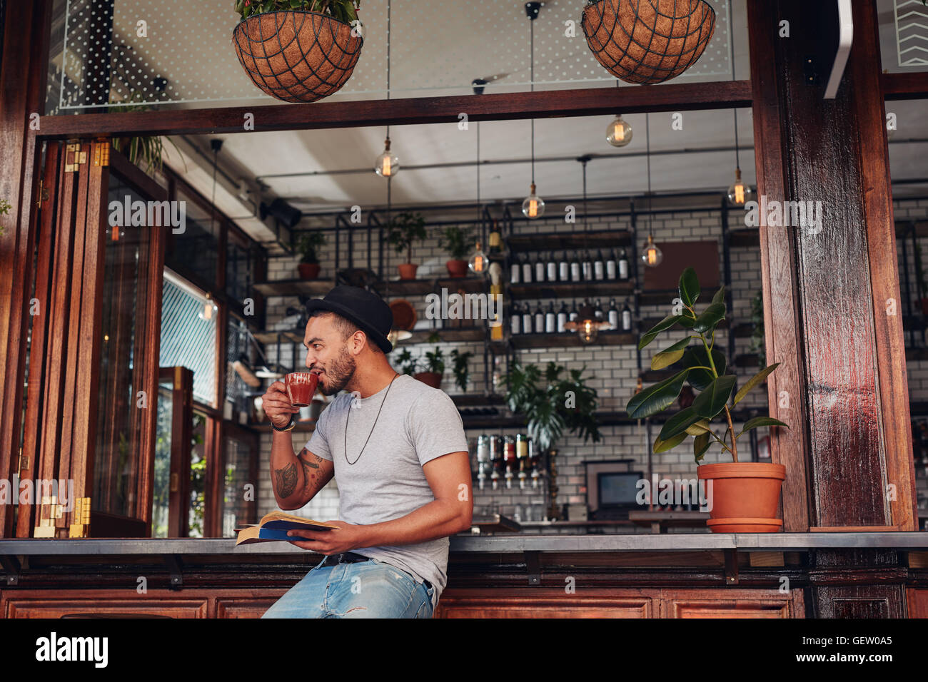 Felice giovane uomo seduto presso il cafe contatore con un libro e di bere il caffè. Elegante maschio caucasico gustando una tazza di caffè a coff Foto Stock