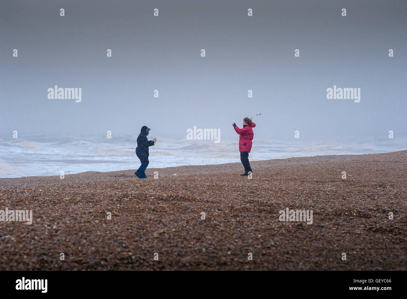 Tempesta di spiaggia femminile, vista di una donna che fotografa la sua amica su una spiaggia deserta e spazzata dal vento a Dunwich, sulla costa del Suffolk, in Inghilterra. Foto Stock