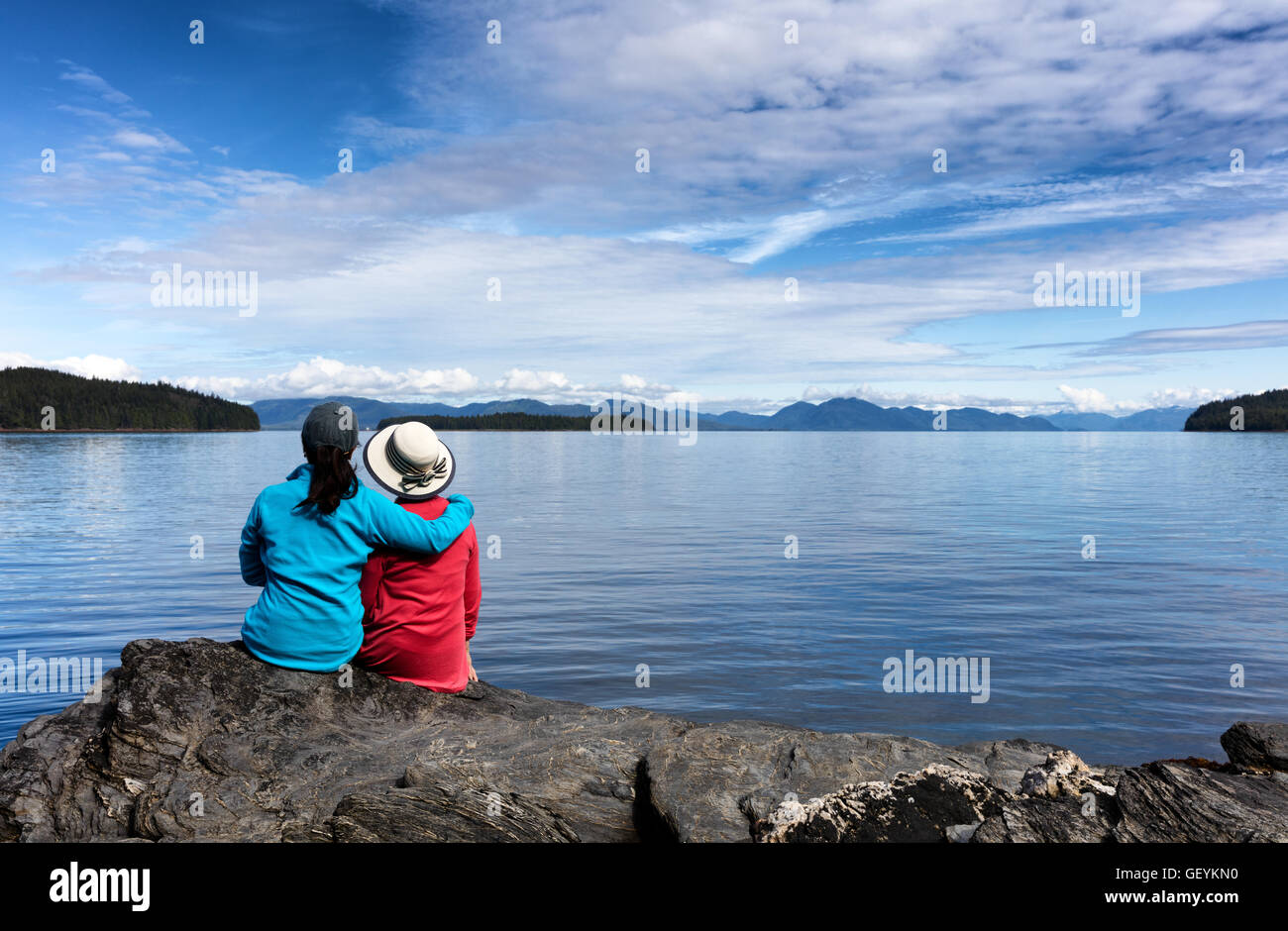 La figlia e la madre, rivolta lontano dalla fotocamera, godendo la vita all'aperto su una bella giornata con il lago e le montagne sullo sfondo. Foto Stock