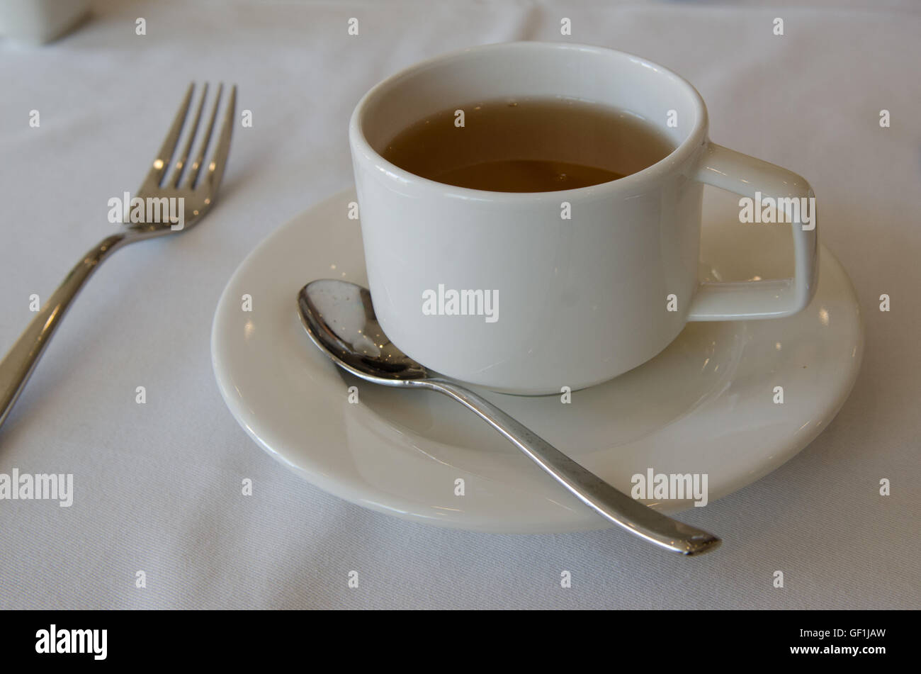 Impostare posto a sedere con bevanda calda nella tazza bianca e il piattino e cucchiaino e la forcella sulla biancheria da letto bianca Foto Stock