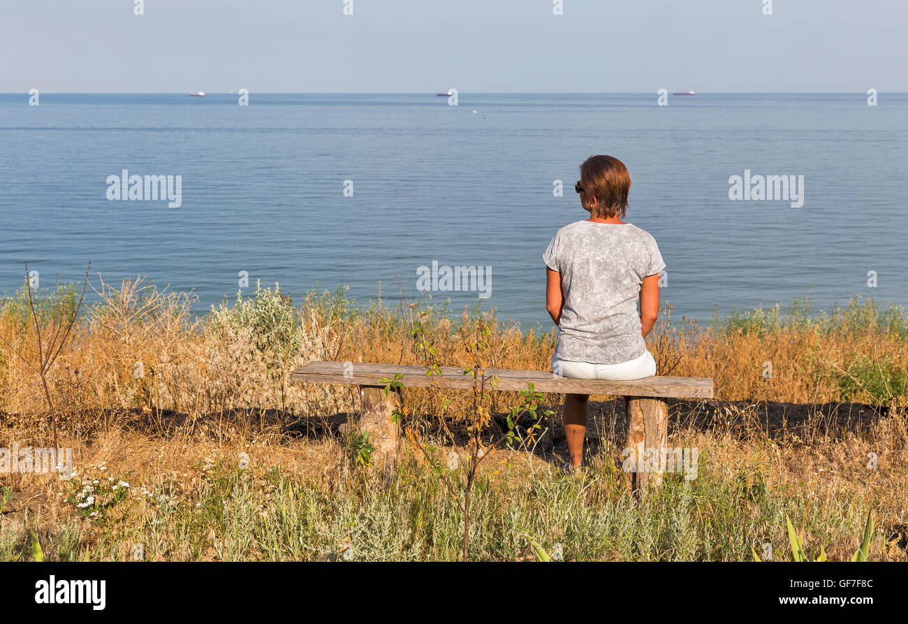 White donna di mezza età con peli più corti in t-shirt, pantaloncini corti e occhiali da sole seduto su una vecchia panca di legno con il Mar Nero seascape Foto Stock