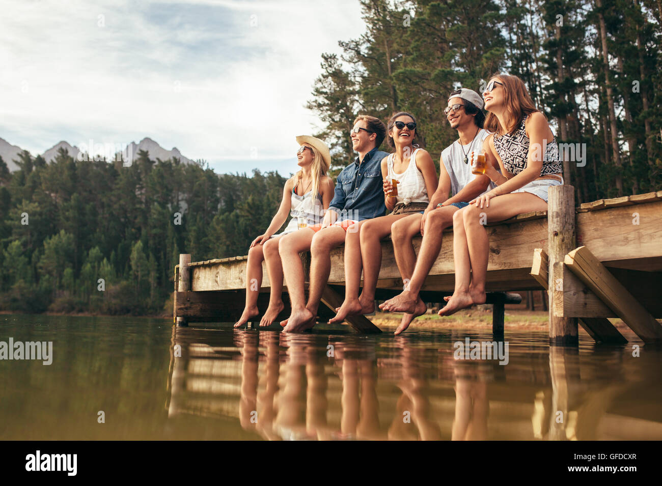Gruppo di felice giovani amici divertendosi e bere birra seduti sul molo al lago. Giovani uomini e donne godendo di un Foto Stock