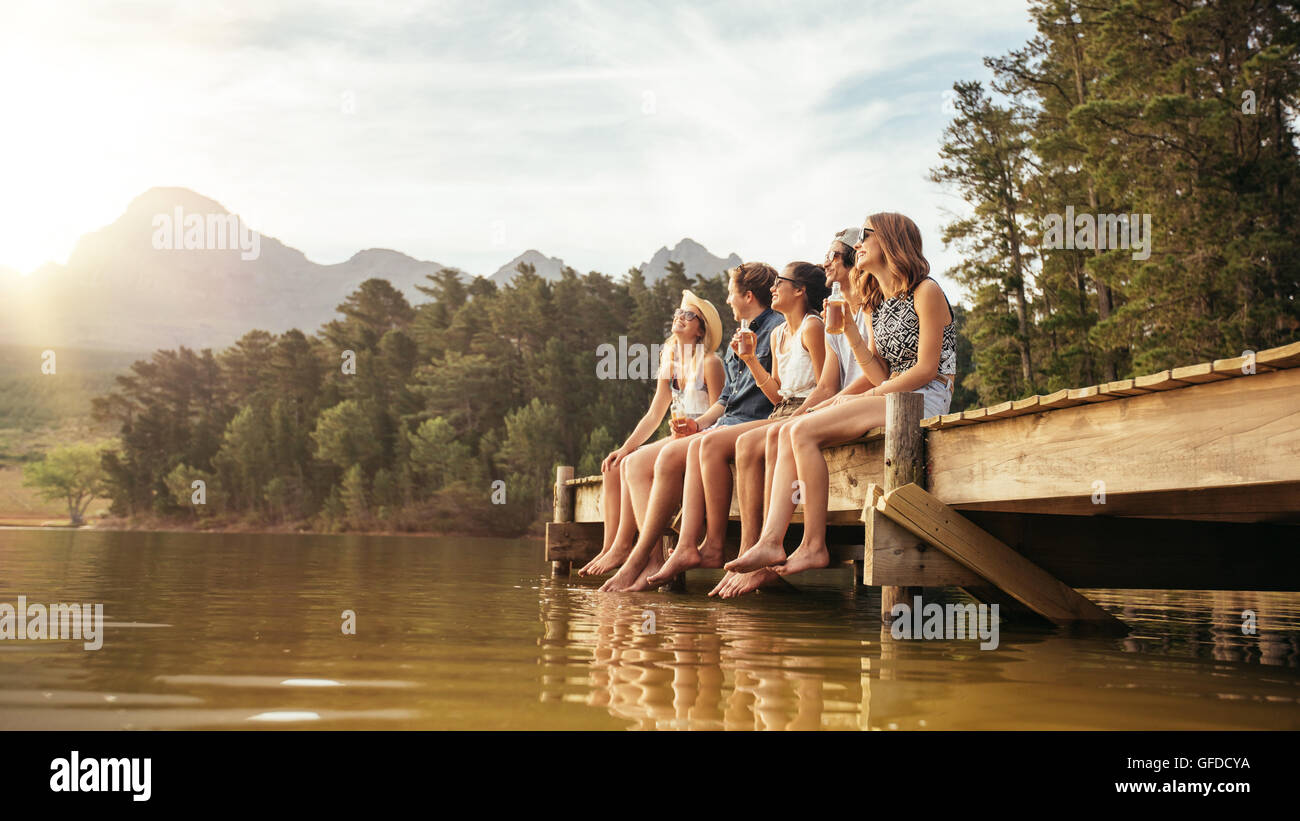 Ritratto di rilassata giovani seduti sul pontile con birre. Amici appendere fuori a lago di un giorno d'estate. Foto Stock