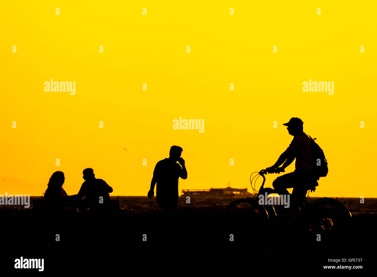 Silhouette di persone di Izmir, Turchia. Andare in bicicletta uomo, un telefono chiamando l'uomo, e due persone seduta su una panchina vicino al mare Foto Stock