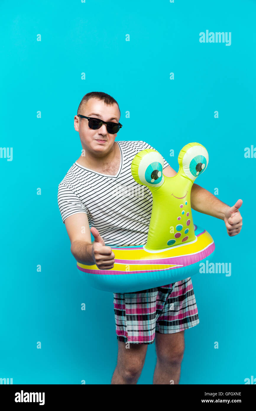Uomo che indossa gli occhiali da sole, maglietta a strisce, nuoto giri in forma di una lumaca guardando la telecamera, pollice in alto, sfondo blu. Concetto di vacanza Foto Stock