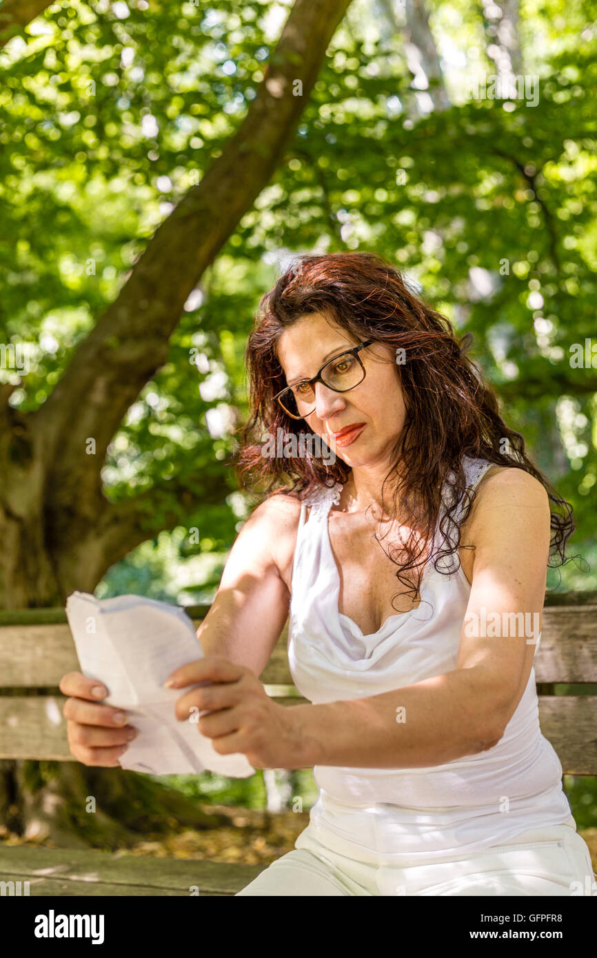 Attraente donna lungimirante con gli occhiali e capelli lunghi legge un usurato foglio di carta su una panchina nel parco tenendolo con le braccia tese Foto Stock