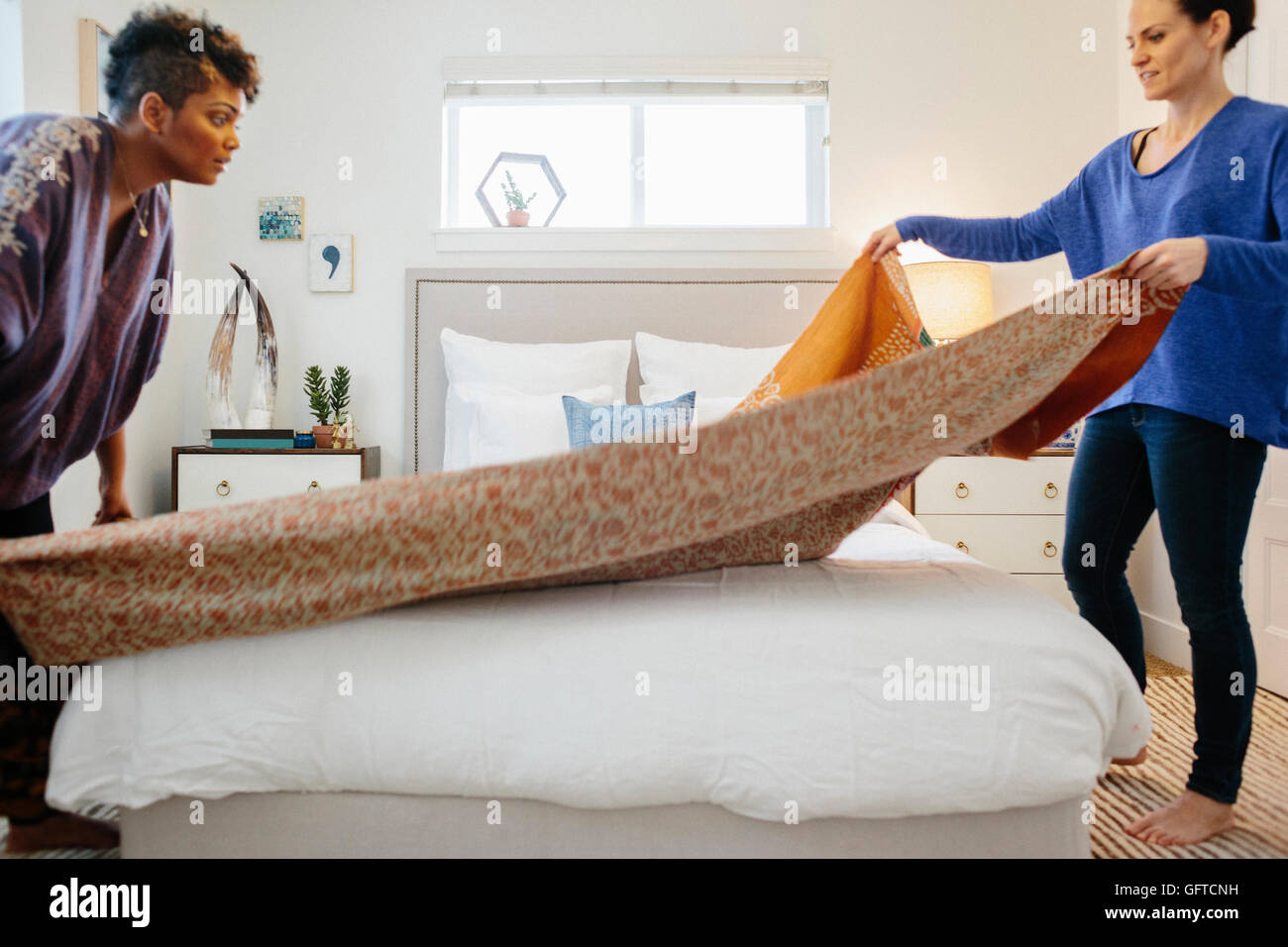 Due donne che compongono un letto diffondendo un quilt modellato sopra il letto matrimoniale in un luminoso e arioso, camera da letto con pareti bianche Foto Stock