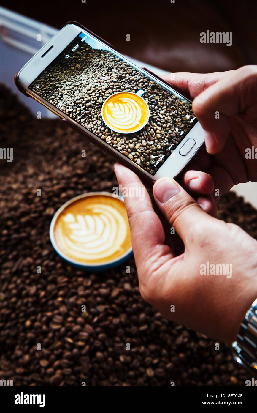 Una persona di scattare una foto con un telefono intelligente di una tazza di caffè sulla sommità di un mucchio di chicchi di caffè tostati Foto Stock