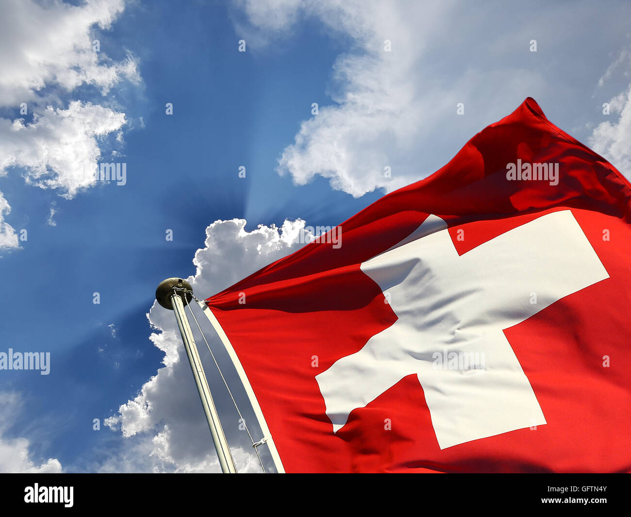 La Svizzera giornata nazionale dello sfondo: bandiera svizzera contro il cielo blu con nuvole bianche e raggi di sole raggiante dietro di essa Foto Stock