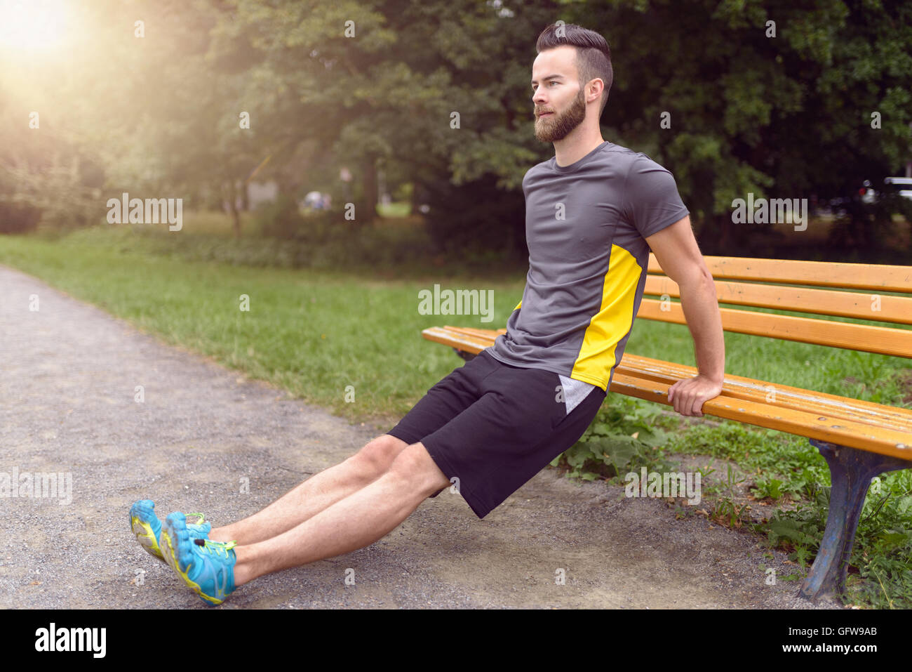 Giovane uomo esercizio utilizzando un di legno una panchina nel parco il sollevamento su se stesso le sue braccia per tonificare i muscoli, vista laterale con copia spazio in una h Foto Stock