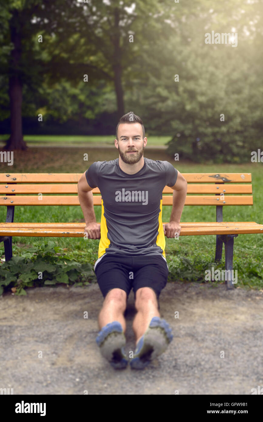 Giovane uomo esercizio utilizzando un di legno una panchina nel parco il sollevamento su se stesso le sue braccia per tonificare i muscoli, in vista frontale con copia spazio in un Foto Stock