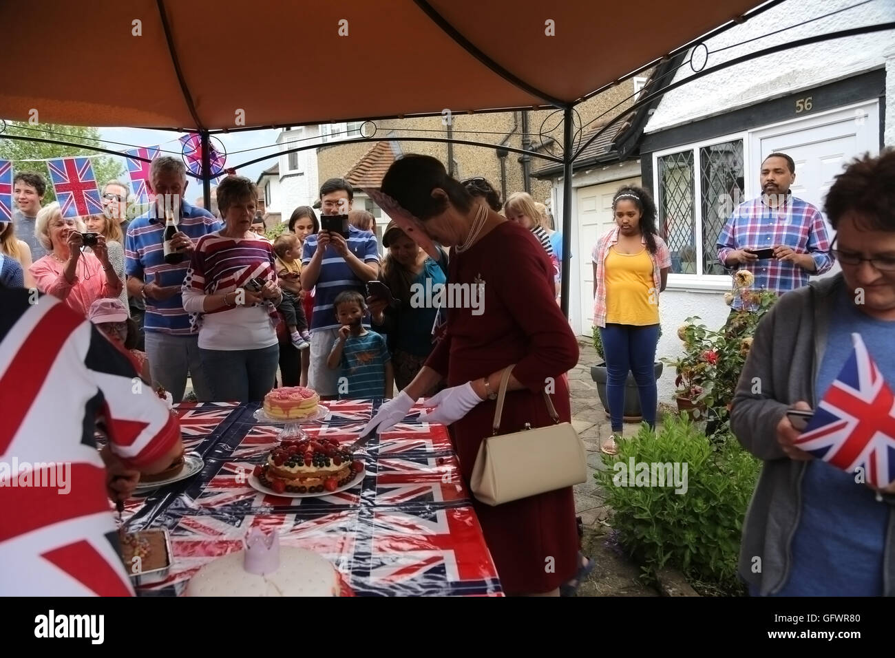 Street partito svoltasi nella celebrazione della Regina Elisabetta II il novantesimo compleanno - donna che indossa una maschera e vestita come la regina tagliare la torta con la gente a guardare la Cornovaglia Road Surrey in Inghilterra Foto Stock