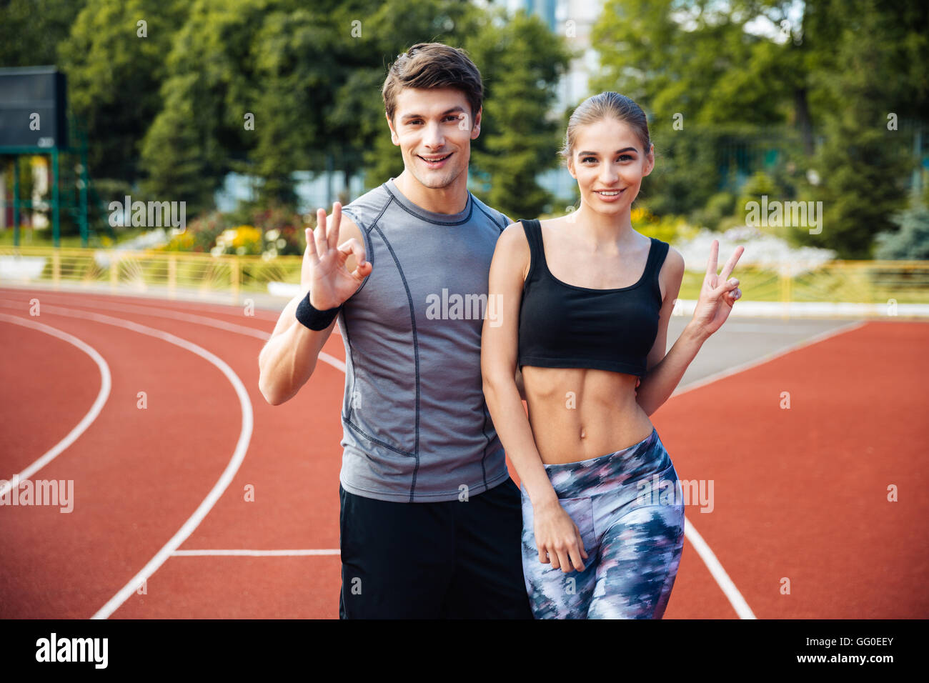 A piena lunghezza shot del giovane uomo e donna in piedi sulla pista di atletica via presso lo stadio che mostra v segno e segno ok Foto Stock