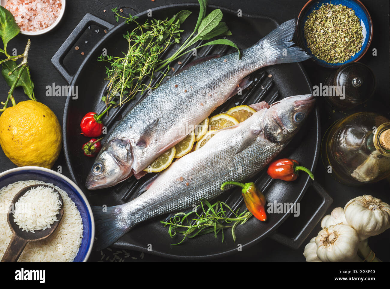 Gli ingredienti per cucinare il pesce sano la cena. Materie non cotti di  spigola con riso, limone, le erbe aromatiche e le spezie sul grill nero  padella di ferro su sfondo scuro