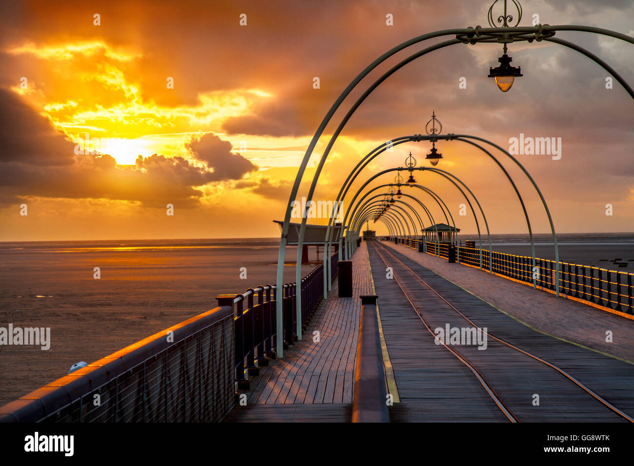Southport, Merseyside, Regno Unito. Il 9 agosto, 2016. Tempesta di scure nuvole sopra il veterano pier struttura dopo la doccia a pioggia, il raffreddamento dell'aria, appena prima del tramonto. Il deep red orb del sole che quasi non si allinea con la riflessione sulle linee del tram creando un illusione di un resort viaggio all'infinito da qualche parte sul mare d'Irlanda costa, con i raggi della sera arrossamento struttura di ferro. Credito: MediaWorldImages/Alamy Live News Foto Stock