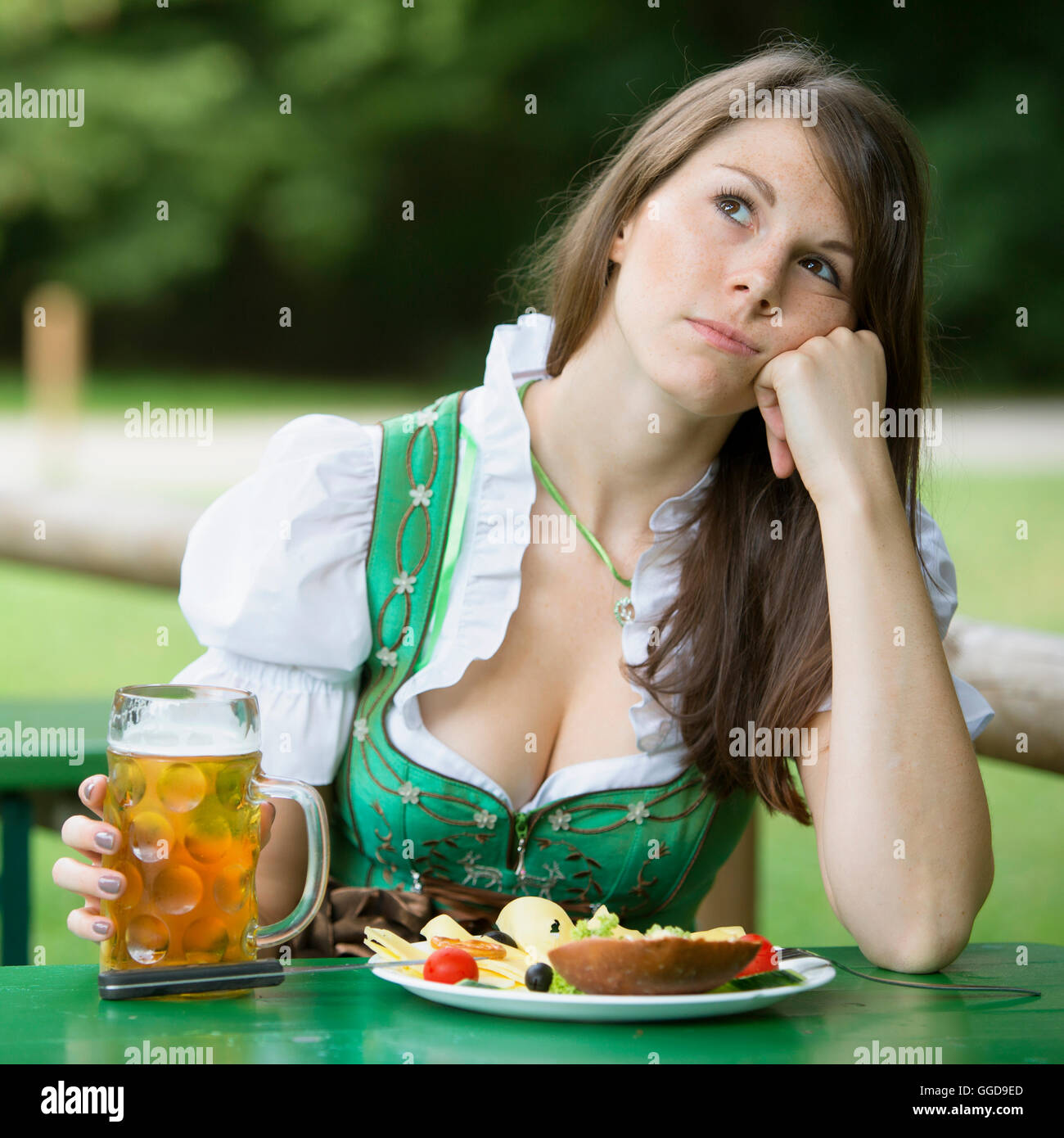 Giovane donna in dirndl seduti al giardino della birra con il cibo e birra e guardando annoiato Foto Stock