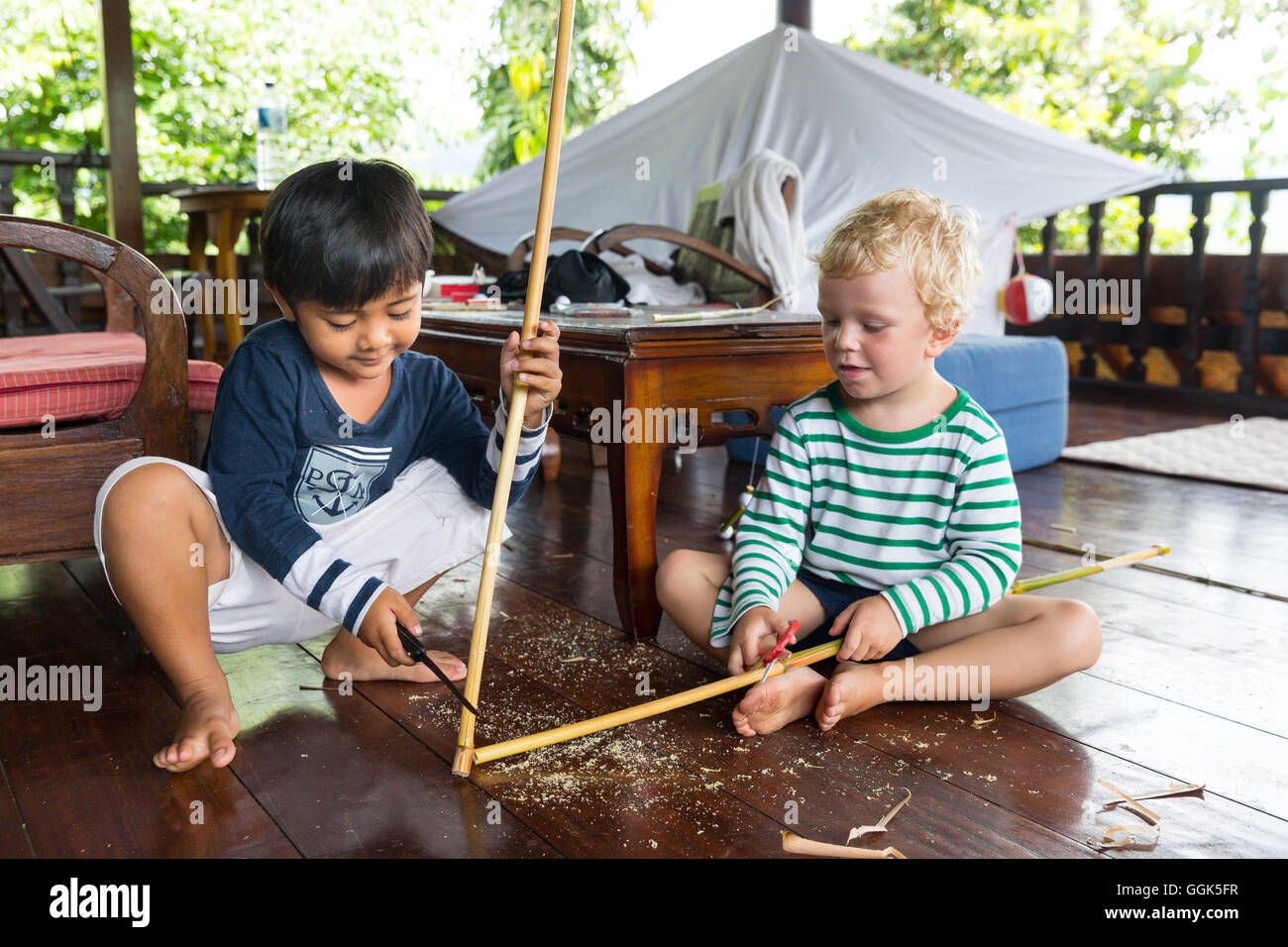 Ragazzo Balinese giocando con il ragazzo straniero, tedesco, 3 anni, bionda, coltello, whittle, carving un bastone di legno, terrazza in legno, Baline Foto Stock