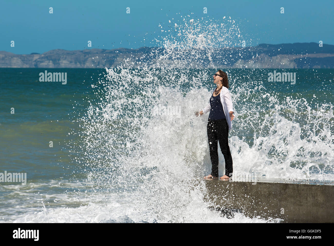 Felice donna in piedi sul mare e ottenere spruzzato da un'onda (MR), Napier, Hawke's Bay, Isola del nord, Nuova Zelanda Foto Stock