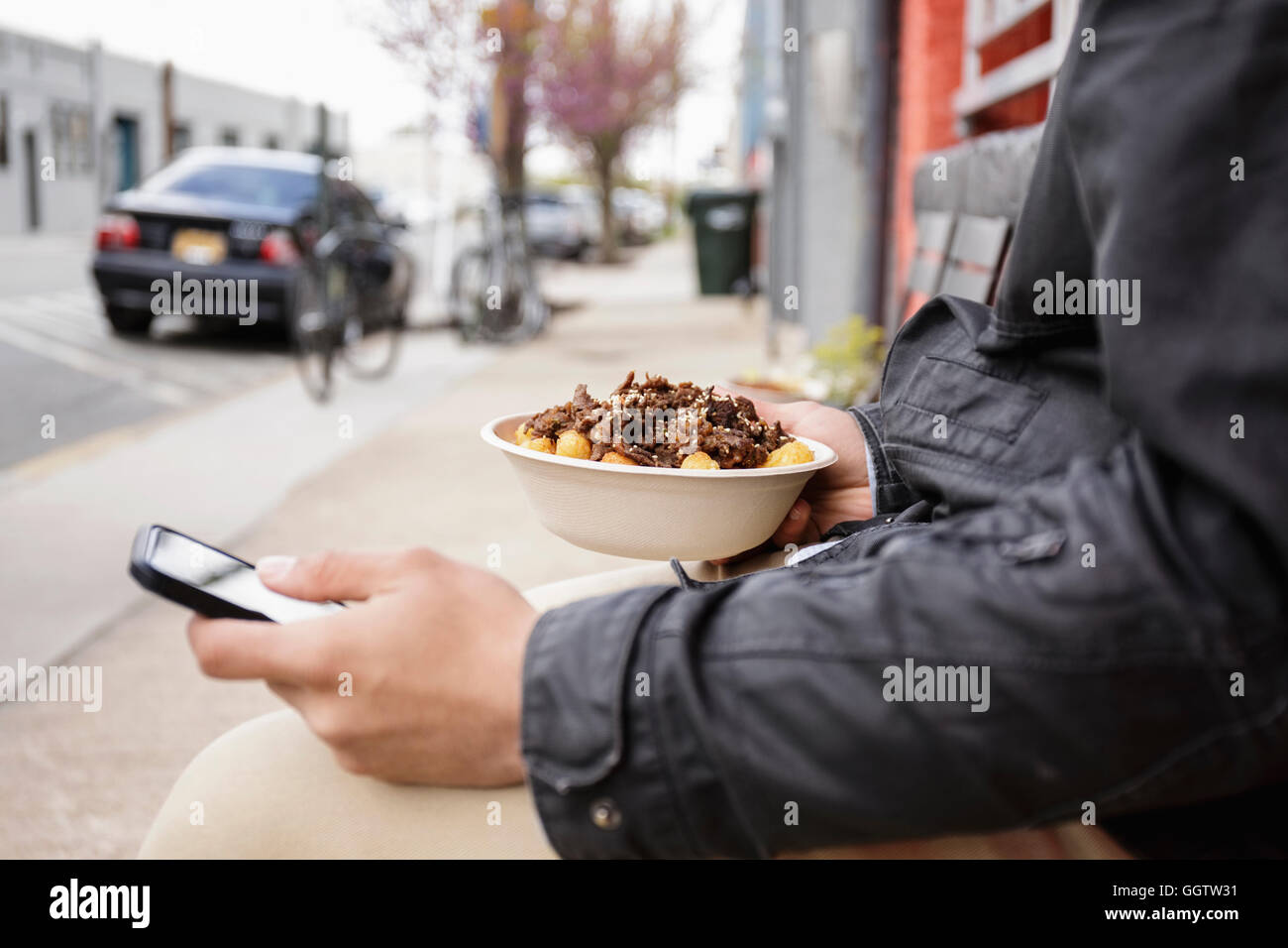 Uomo ispanico mangiare una ciotola di cibo sul marciapiede tramite telefono cellulare Foto Stock