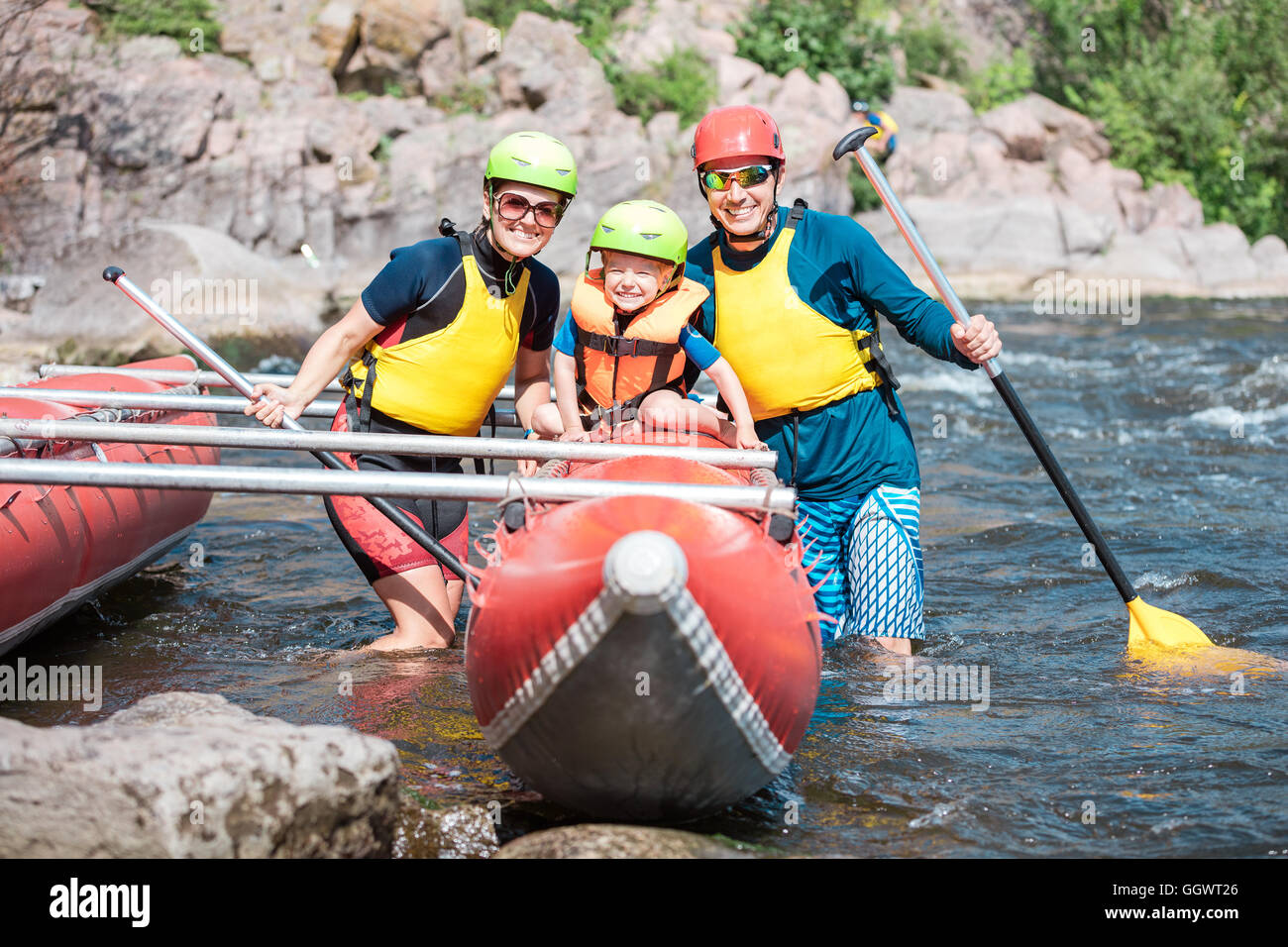 Giovane famiglia di tre indossare giubbotti salvagente e caschi in posa accanto a catamarano a remi Foto Stock