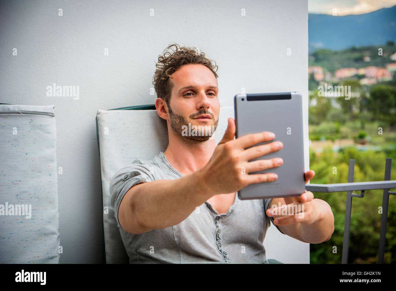 Bello alla moda uomo che indossa la t-shirt seduto e facendo la videochat, guardando verso il basso in corrispondenza di un computer tablet che egli è in possesso, all'aperto o Foto Stock