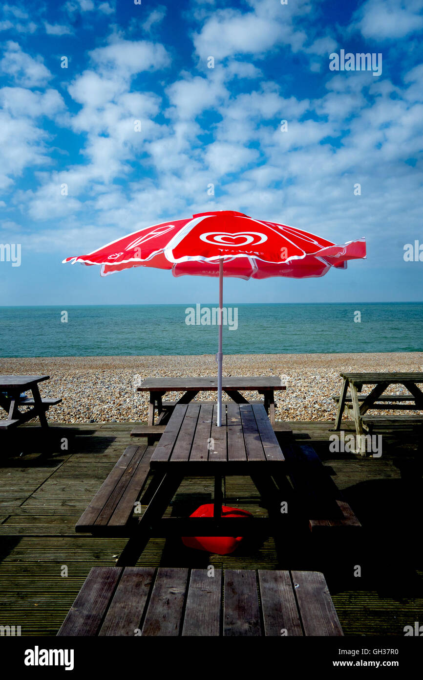 Red & White ombrello sul tavolo in legno e panchine in Seaside Cafe, il mare e la spiaggia di background, cielo blu con nuvole bianche Foto Stock