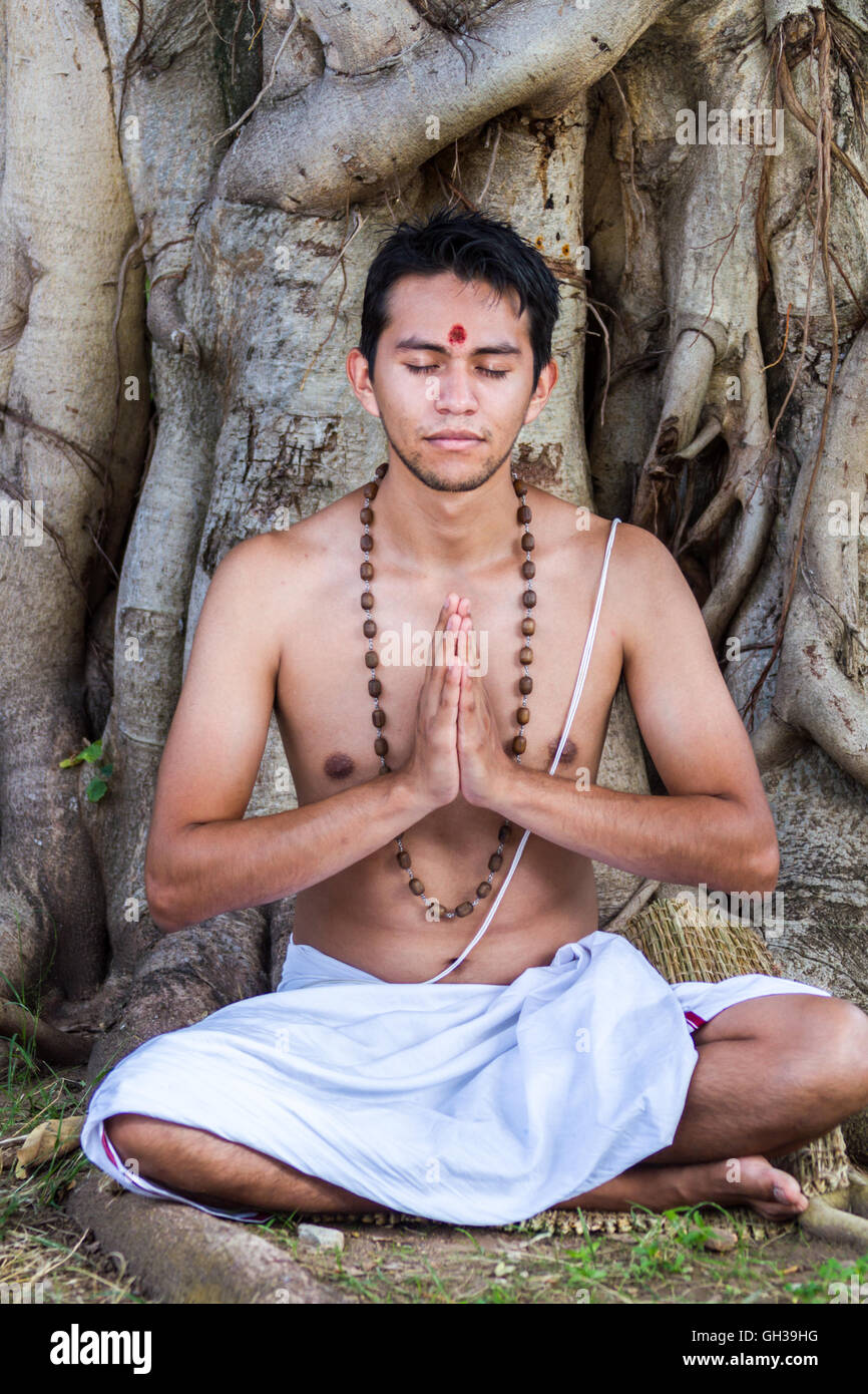 Un giovane uomo indù si siede in meditazione sotto un banyan tree. Foto Stock