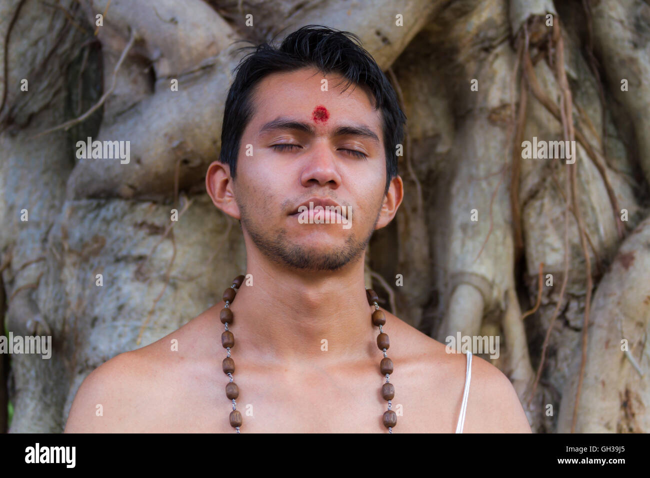 Un giovane uomo indù si siede in meditazione sotto un banyan tree. Foto Stock