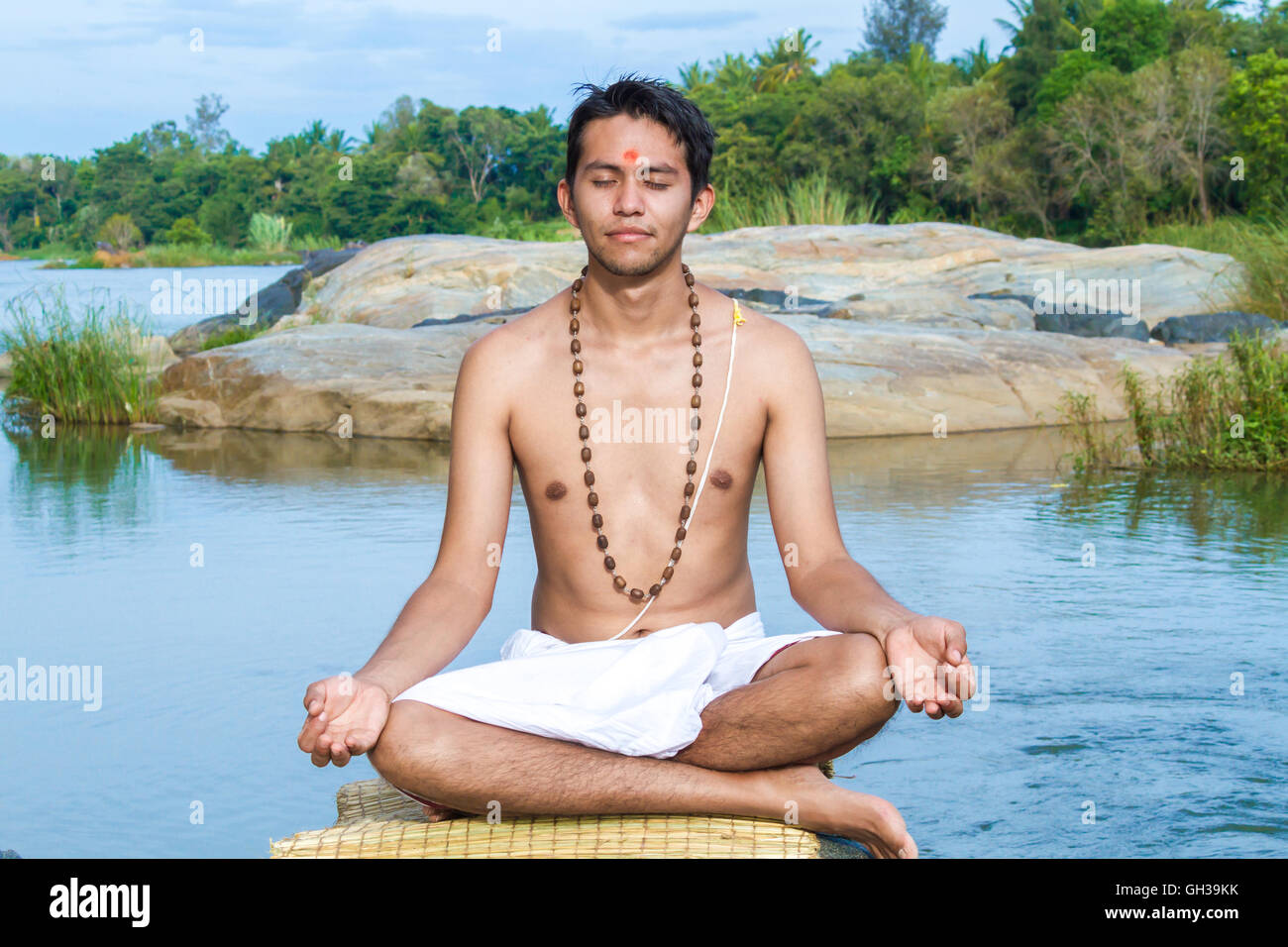 Un giovane sacerdote Indù (bramino), siede in meditazione su una riva di un fiume. Foto Stock