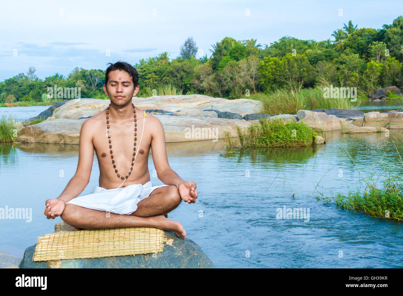 Un giovane sacerdote Indù (bramino), siede in meditazione su una riva di un fiume. Foto Stock