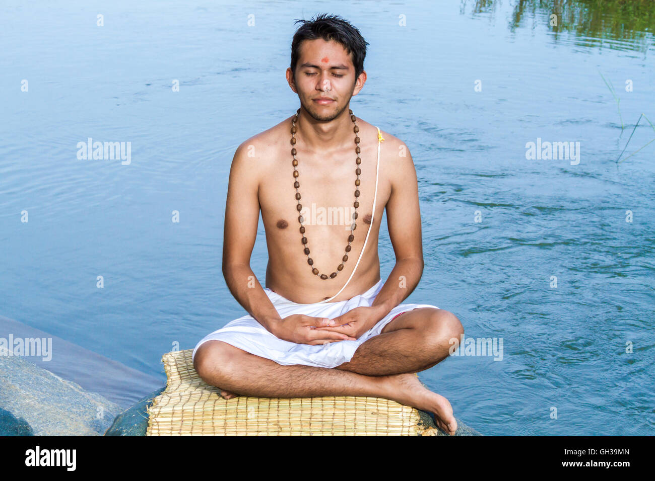 Un giovane sacerdote Indù (bramino), siede in meditazione su una riva di un fiume. Foto Stock