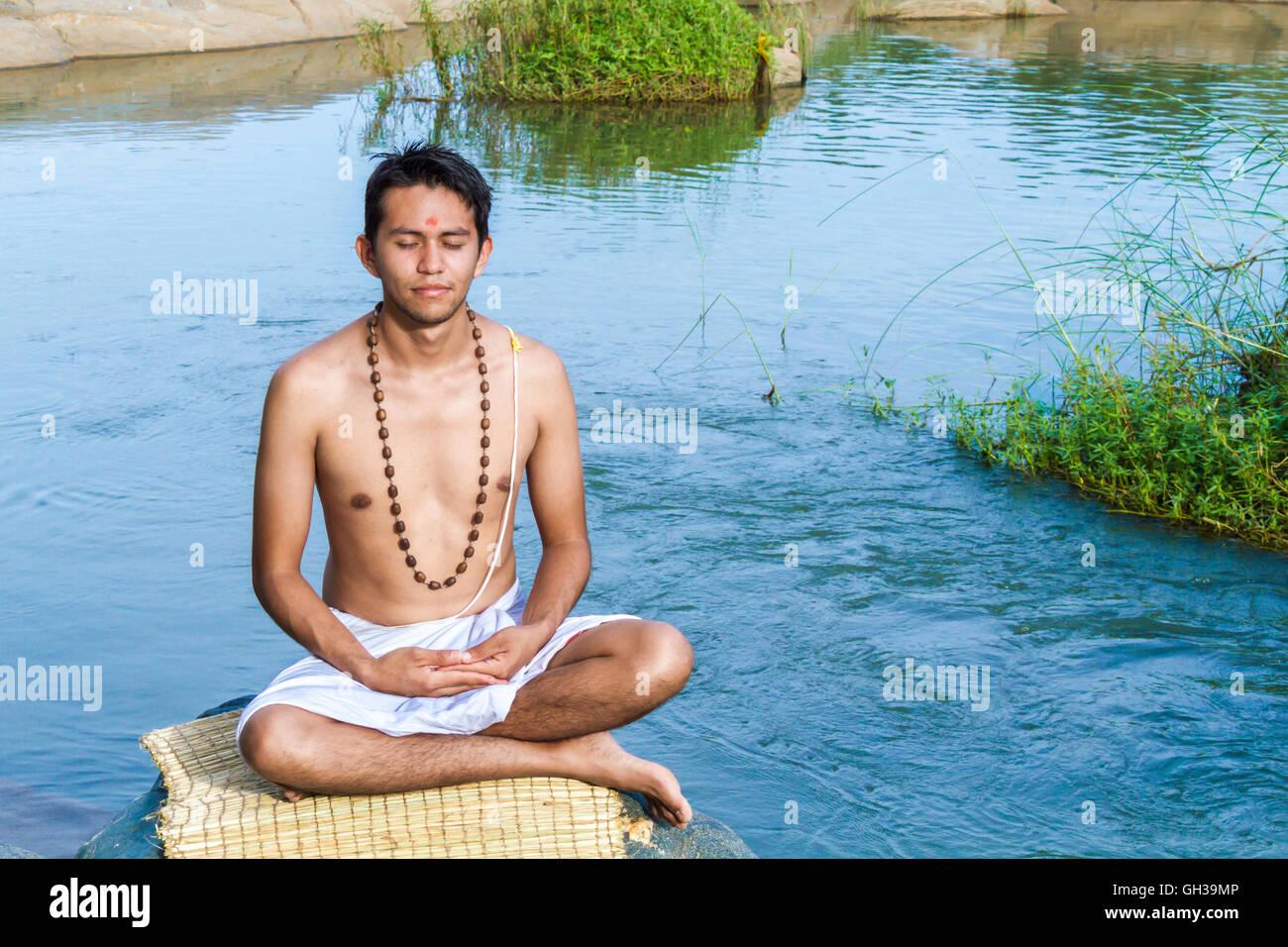 Un giovane sacerdote Indù (bramino), siede in meditazione su una riva di un fiume. Foto Stock