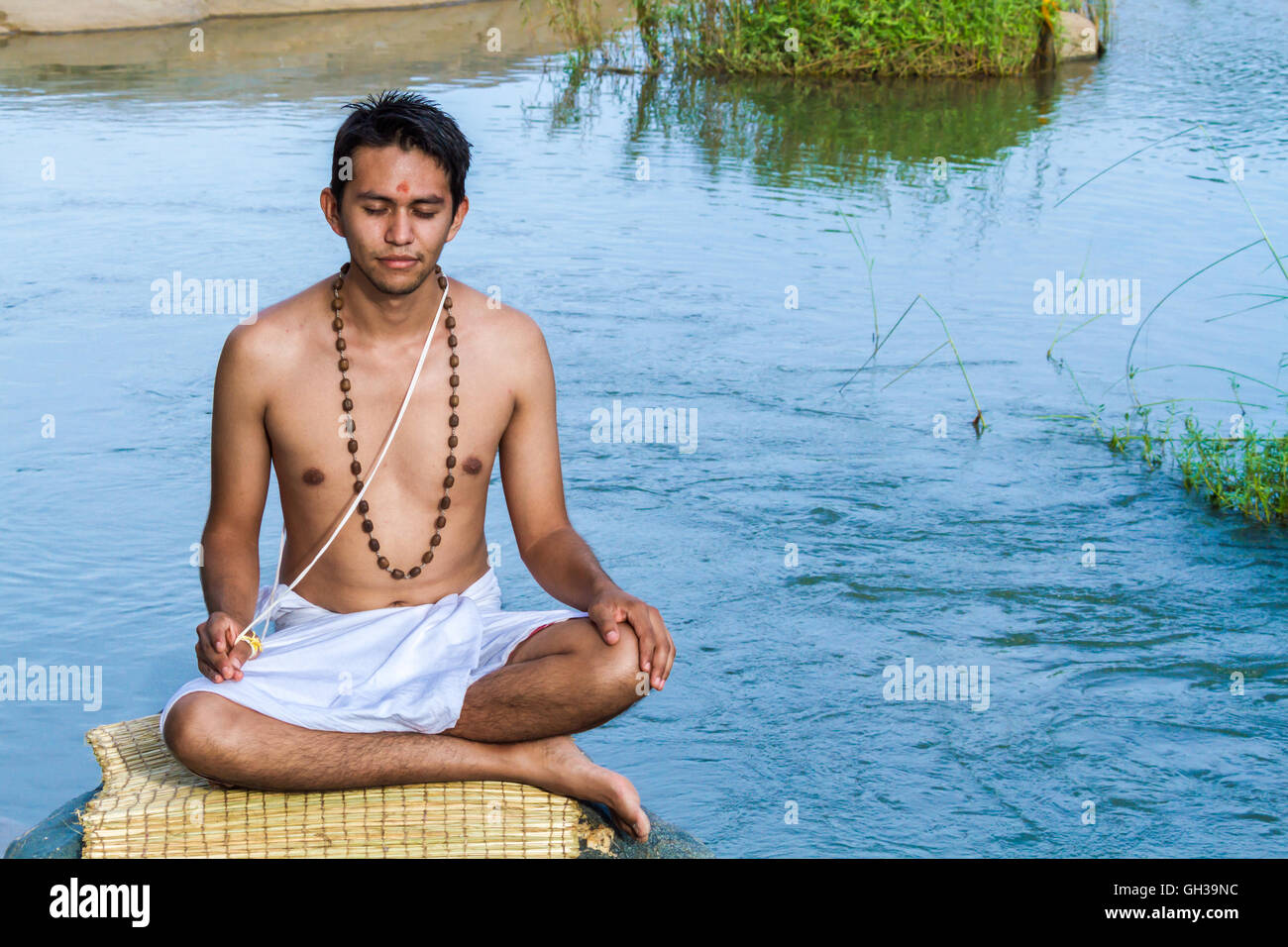 Un giovane sacerdote Indù (bramino) si siede in meditazione su una riva di un fiume. Foto Stock