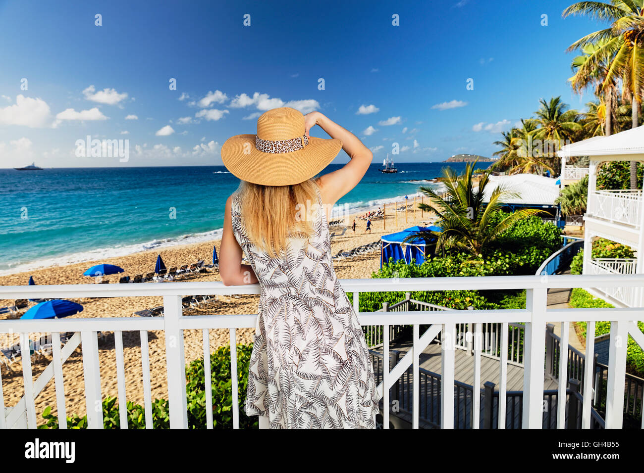 Vista posteriore di una donna in abiti estivi e Hat guardando fuori di un resort sulla spiaggia da un balcone Foto Stock