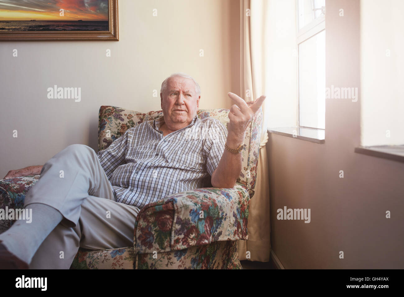 Piscina colpo di un senior caucasica uomo seduto a assisted living facility. Uomo anziano seduto su una sedia di braccio dalla finestra a Foto Stock